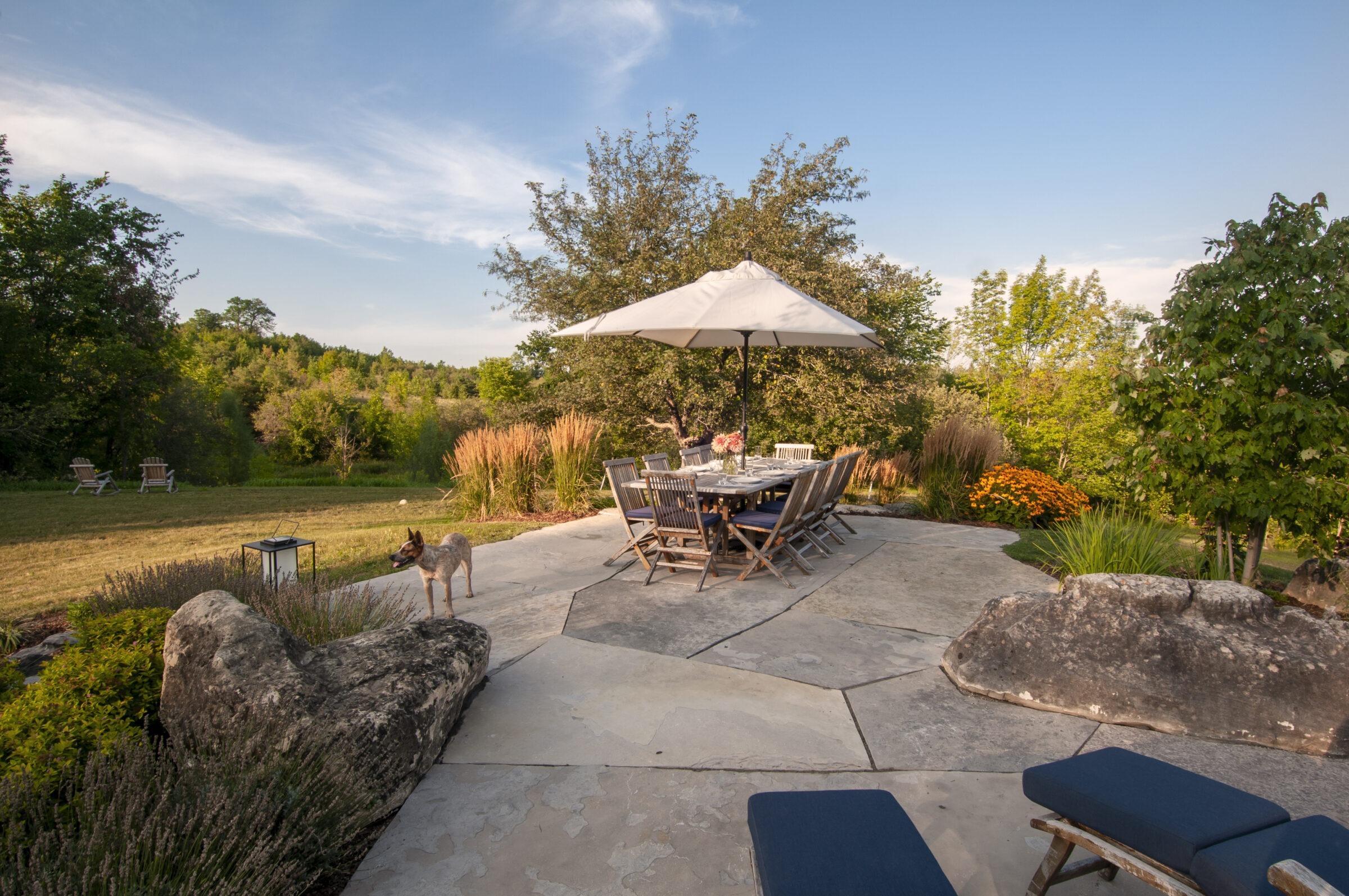 A tranquil patio with outdoor dining setup, surrounded by greenery and flowers, features a dog and several chairs on the lawn.