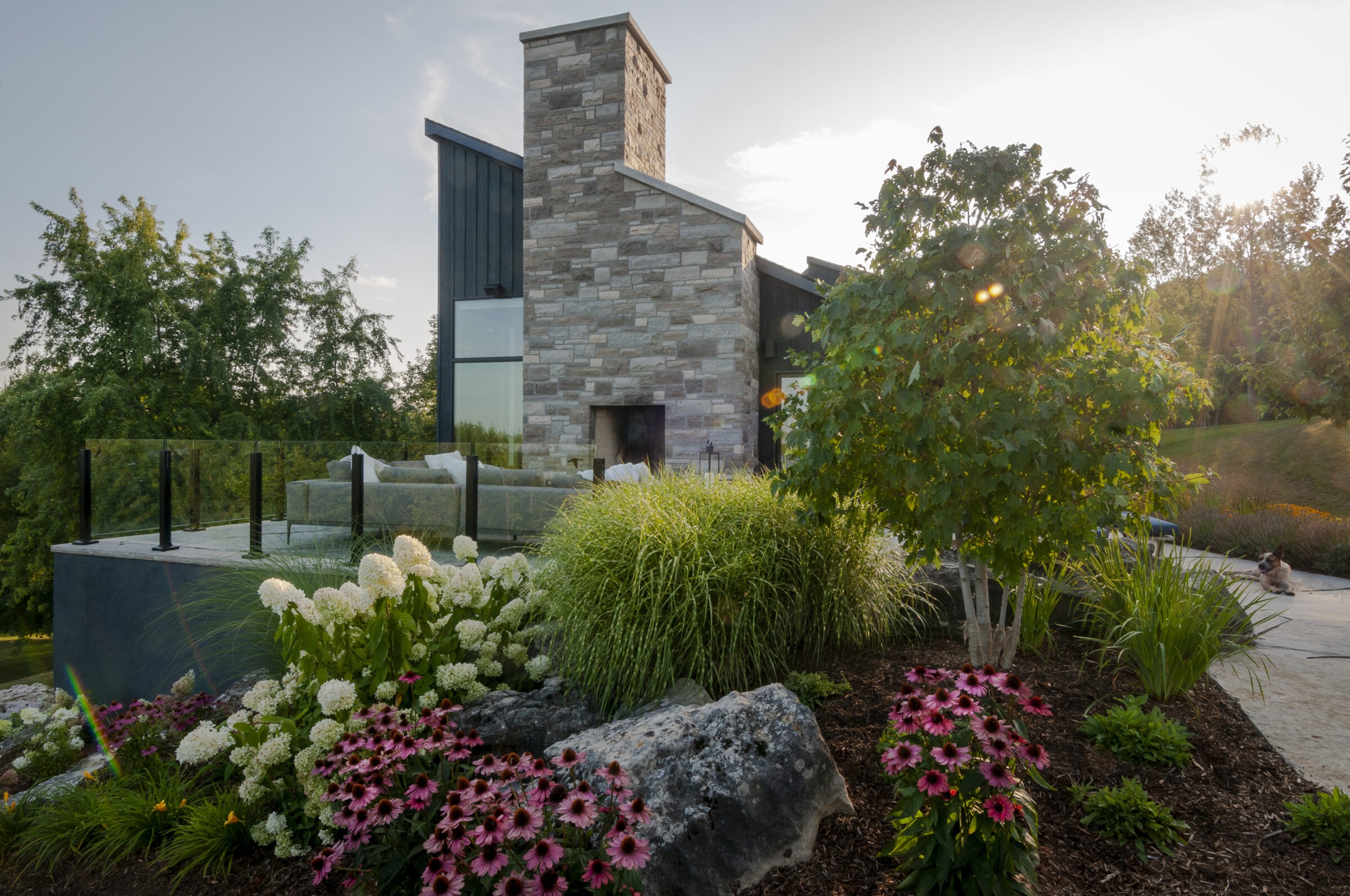 Modern house with stone chimney and glass railing, surrounded by lush garden featuring pink and white flowers. Sunlight filters through the trees.