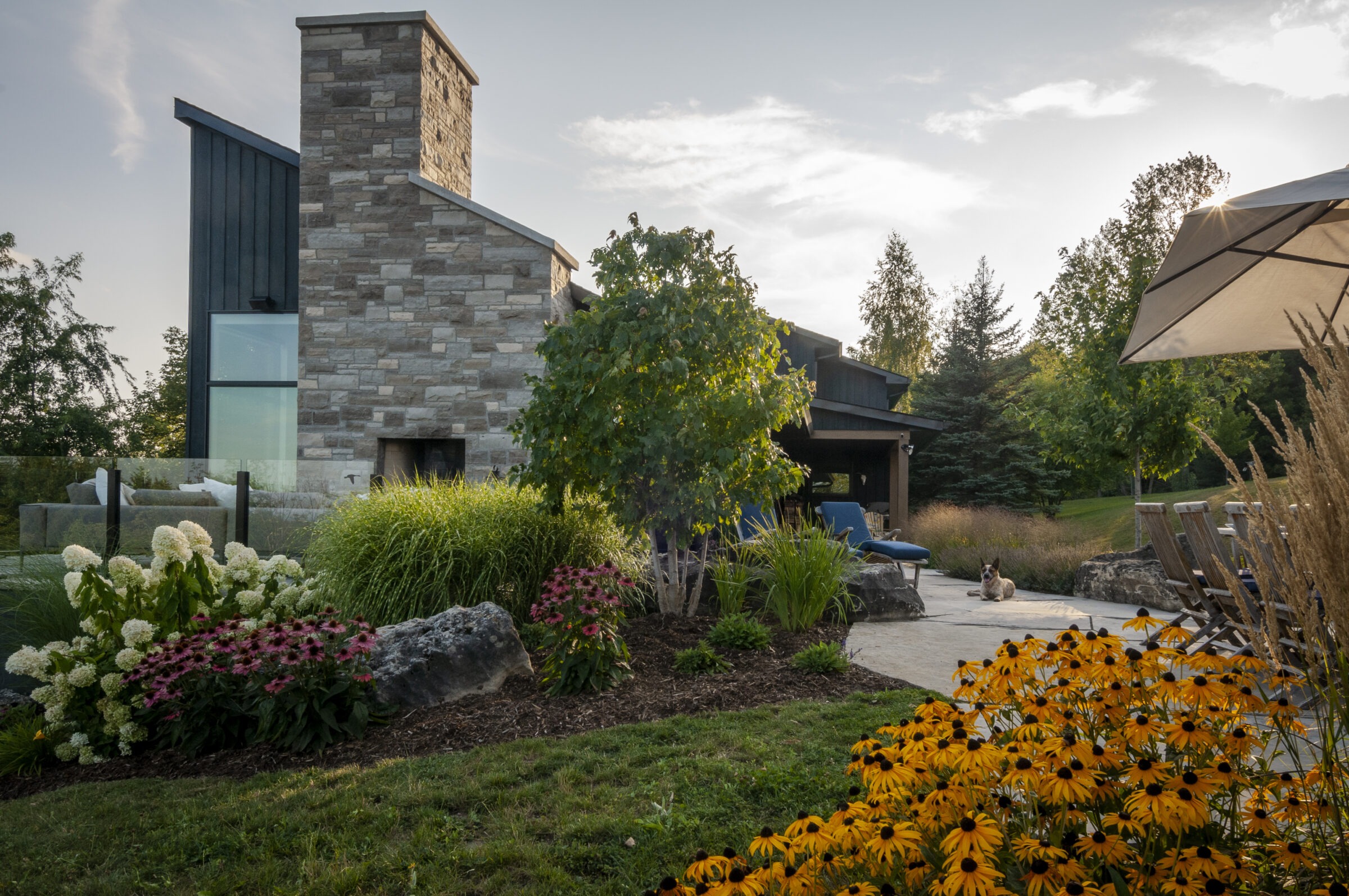 Modern stone house with lush garden, patio area, and a dog relaxing. Peaceful, sunlit scene surrounded by trees and vibrant flowers.