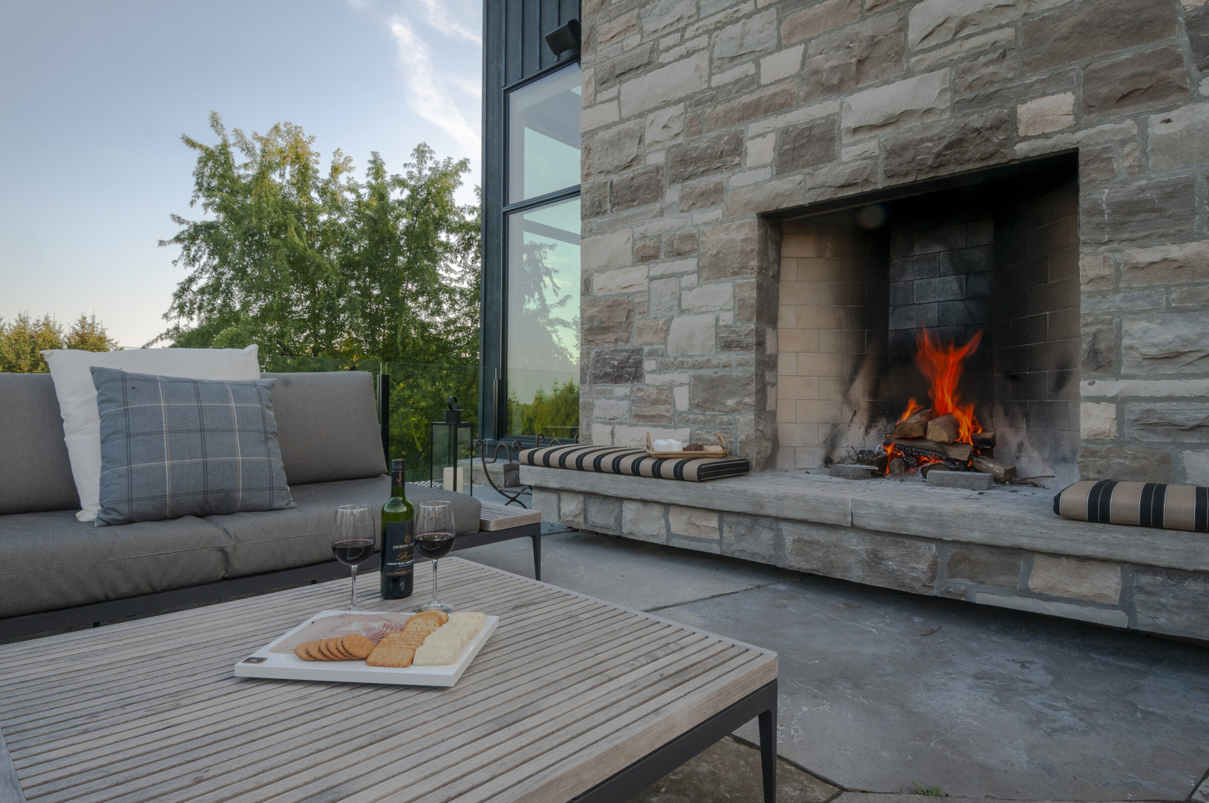 Outdoor patio featuring a cozy fireplace, comfortable seating, and a table with wine and snacks. Lush greenery provides a serene backdrop.