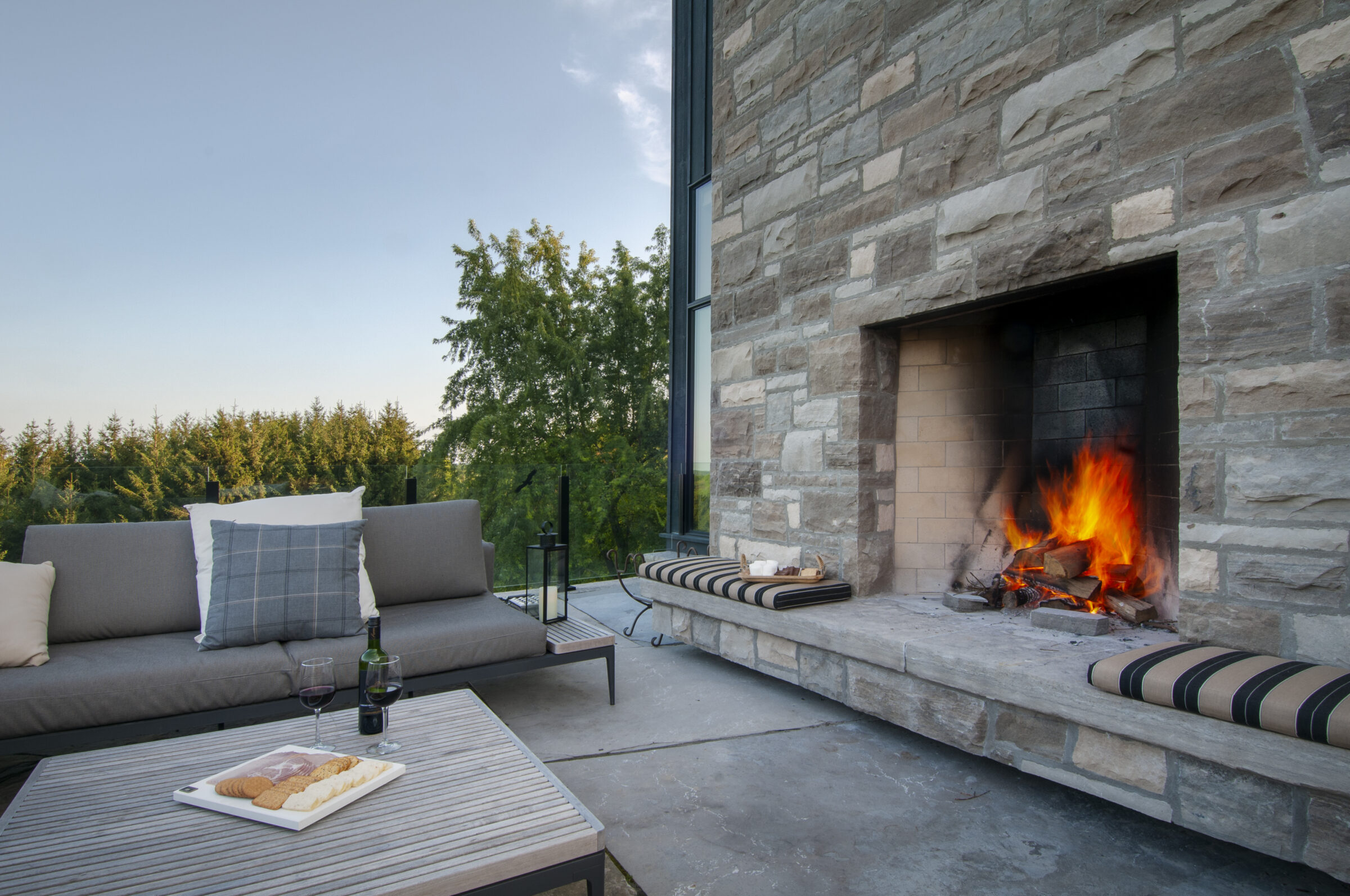 Outdoor patio with stone fireplace, burning fire, comfortable seating, and a table with wine and snacks, set against a backdrop of trees.