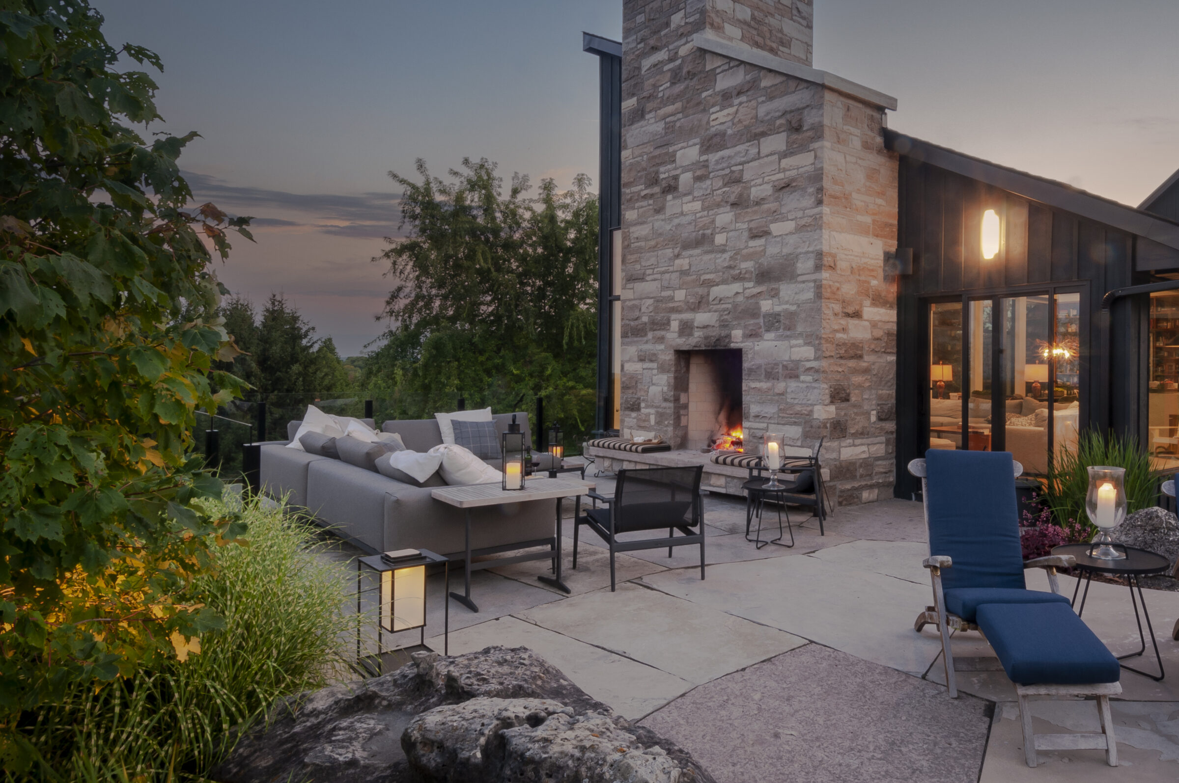 Modern patio with stone fireplace, comfortable seating, and ambient lighting. Evening setting surrounded by greenery, creating a cozy outdoor retreat.