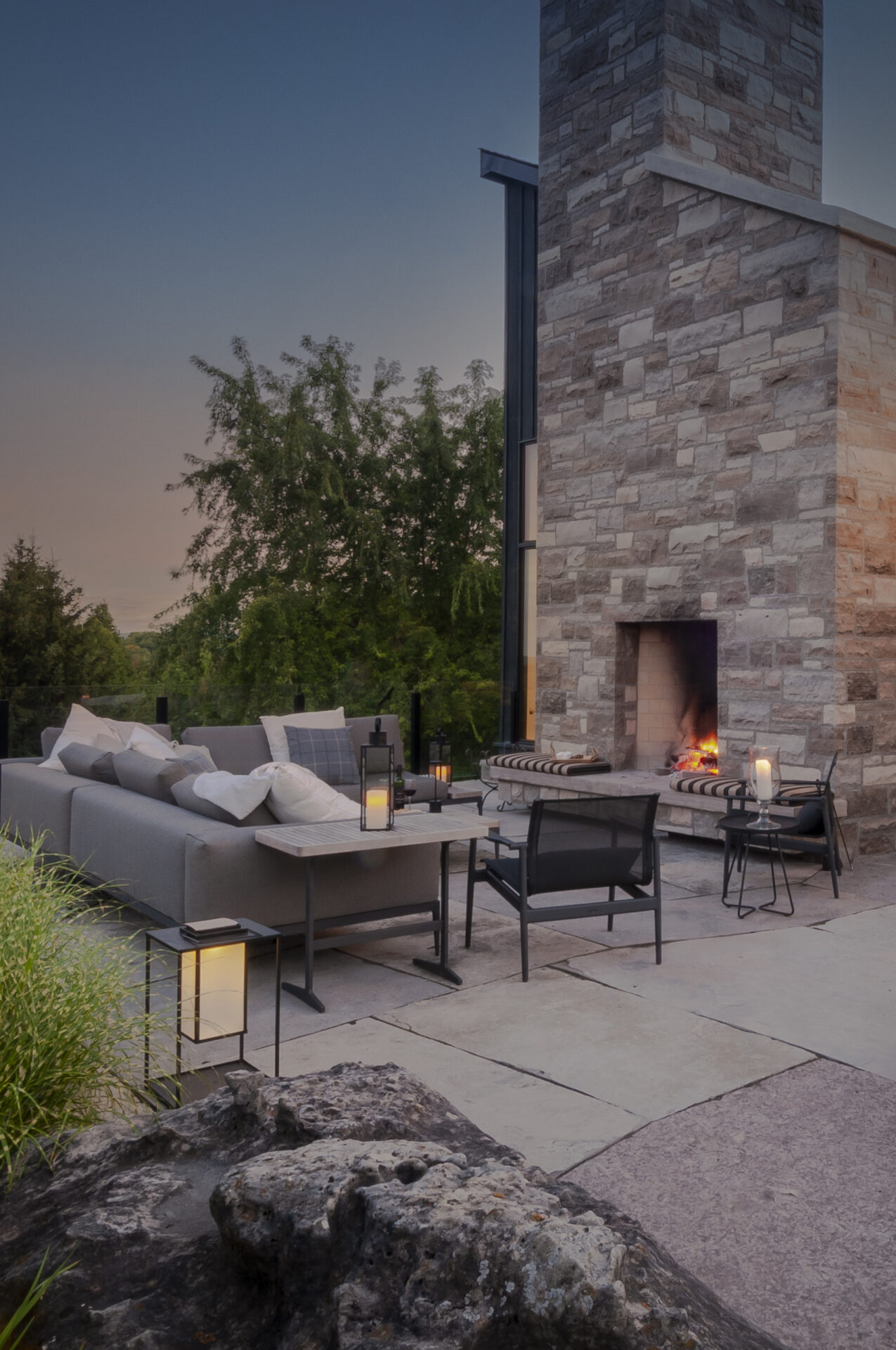 A cozy outdoor patio features modern furniture, a stone fireplace, and soft lighting surrounded by lush greenery at dusk.