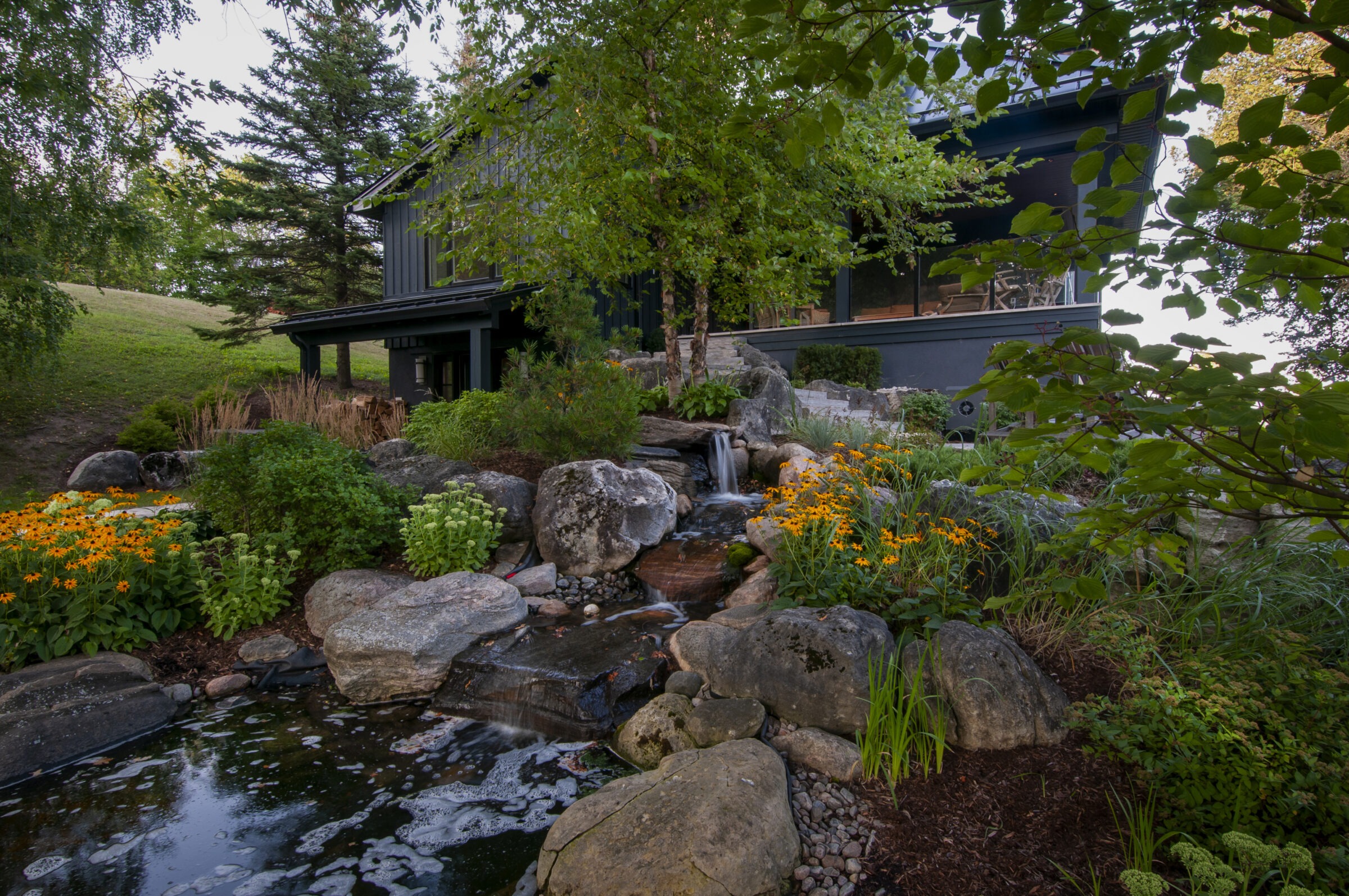 A modern house surrounded by lush greenery, colorful flowers, rocks, and a small waterfall cascading into a tranquil pond.