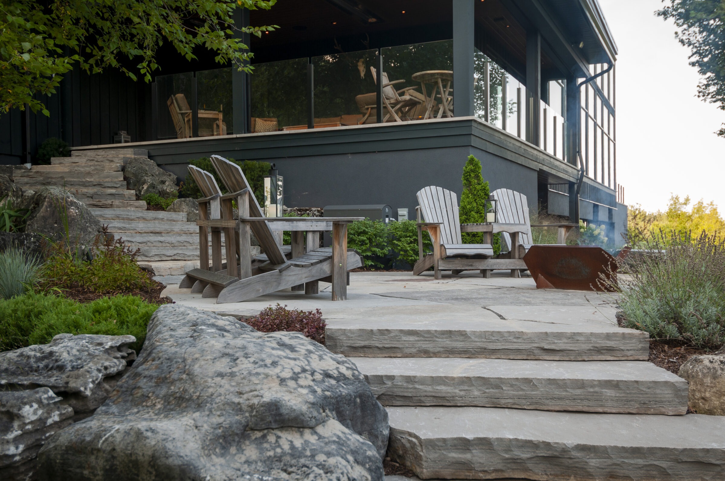 Modern patio with Adirondack chairs, fire pit, and stone steps leading to a glass-railed veranda, surrounded by lush vegetation and large rocks.