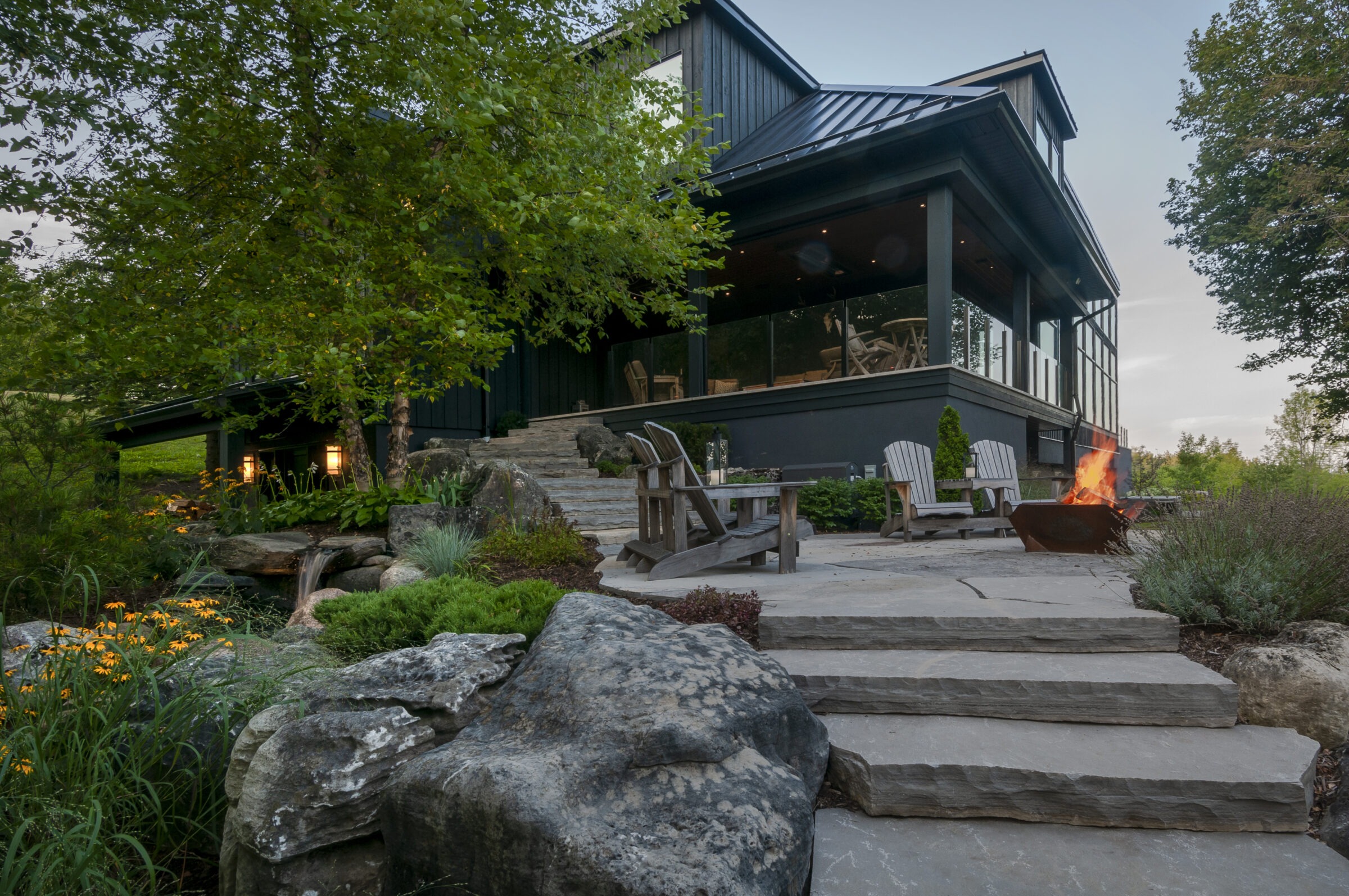 A modern house with a metal roof, surrounded by lush greenery, stone steps, and a cozy patio with a fire pit and chairs.