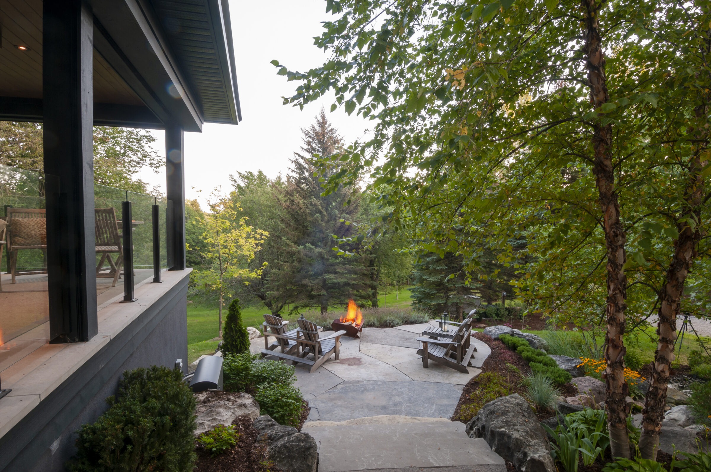 Charming garden scene with fire pit, surrounded by Adirondack chairs and trees. A cozy porch overlooks the lush, serene landscape.