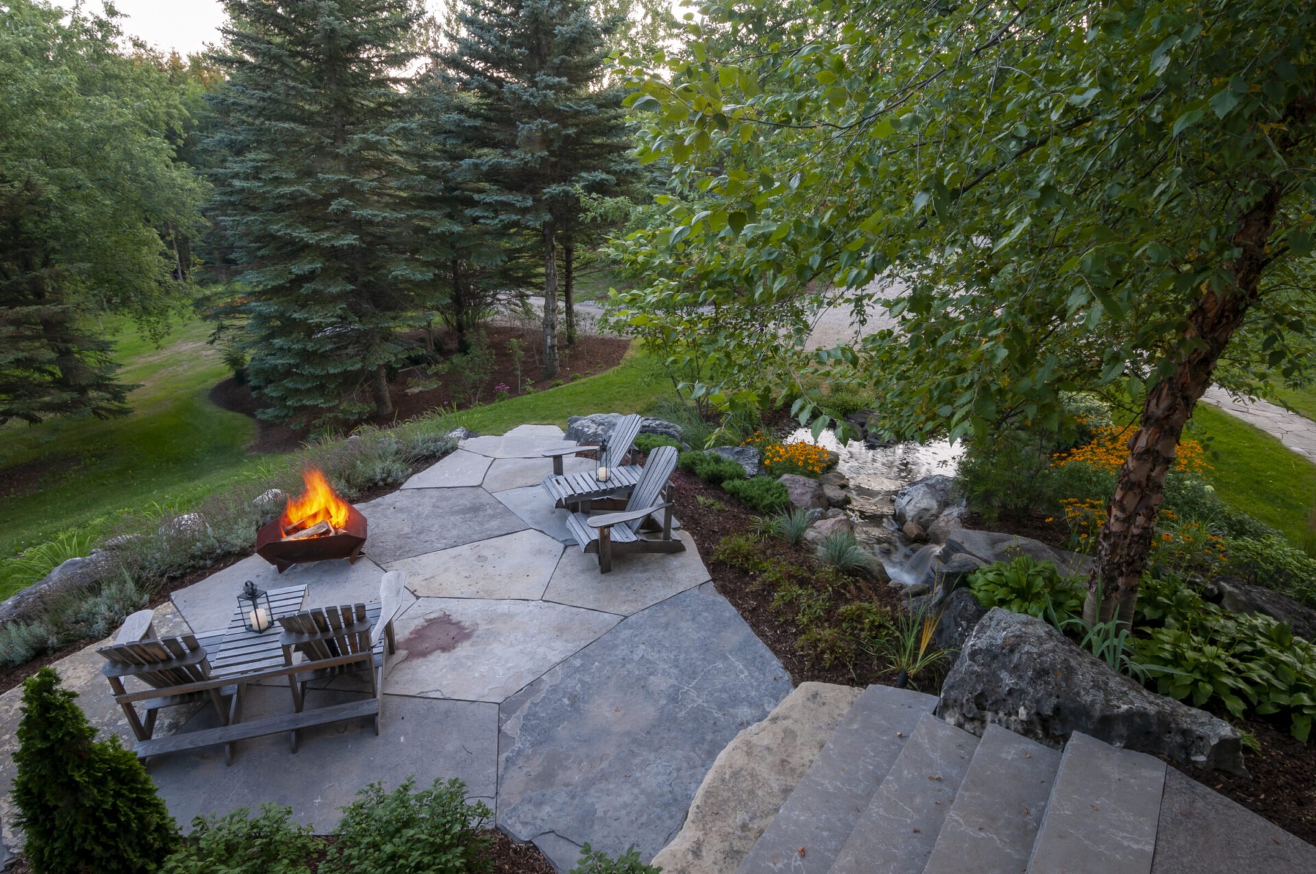 A tranquil garden with a stone patio features a lit fire pit, wooden chairs, and lush green surroundings.