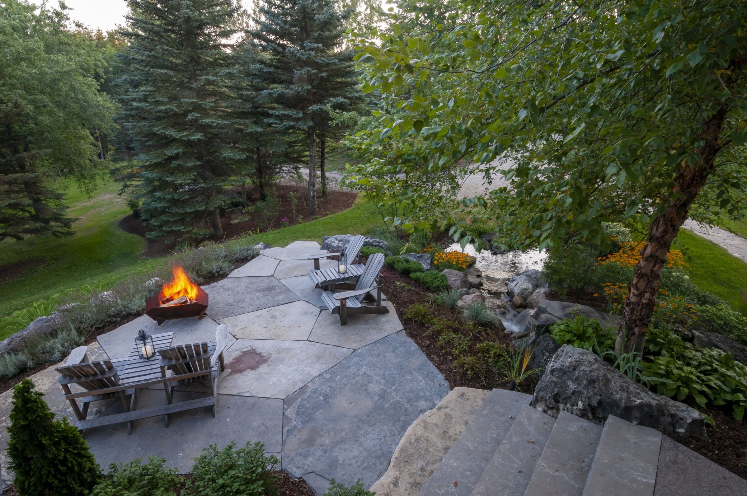 A tranquil garden with a stone patio features a lit fire pit, wooden chairs, and lush green surroundings.