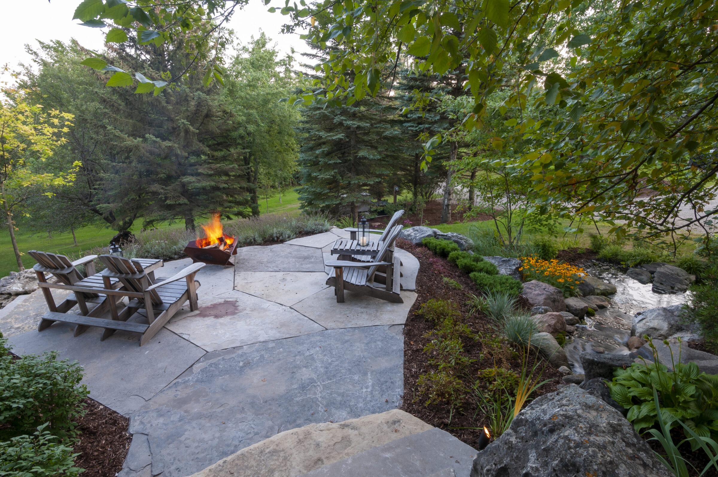 A serene garden patio with Adirondack chairs around a fire pit, surrounded by lush greenery, rocks, and a small stream.