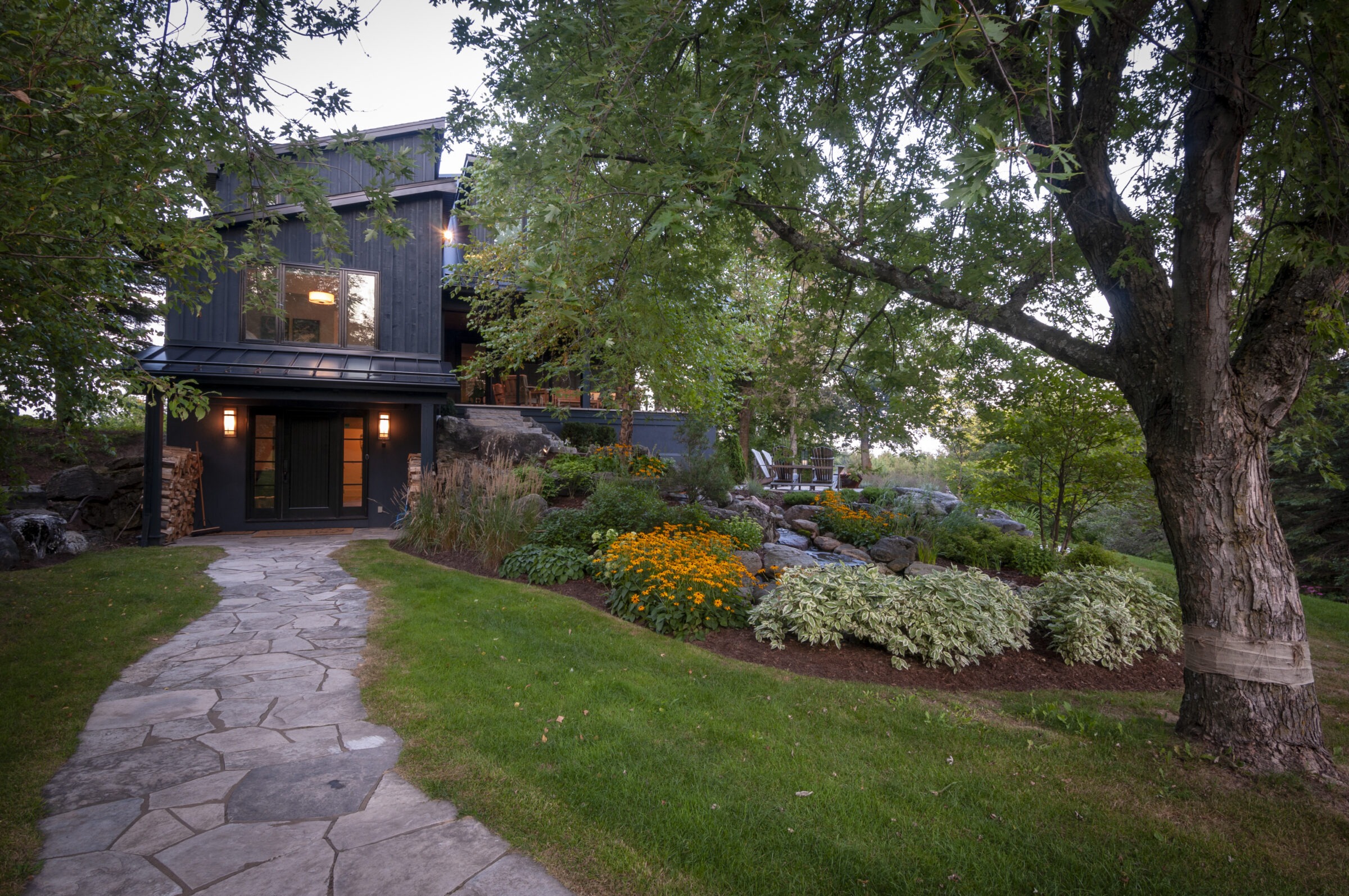 Modern house surrounded by lush greenery with stone path, vibrant flowers, and large trees creating a serene, inviting outdoor space.
