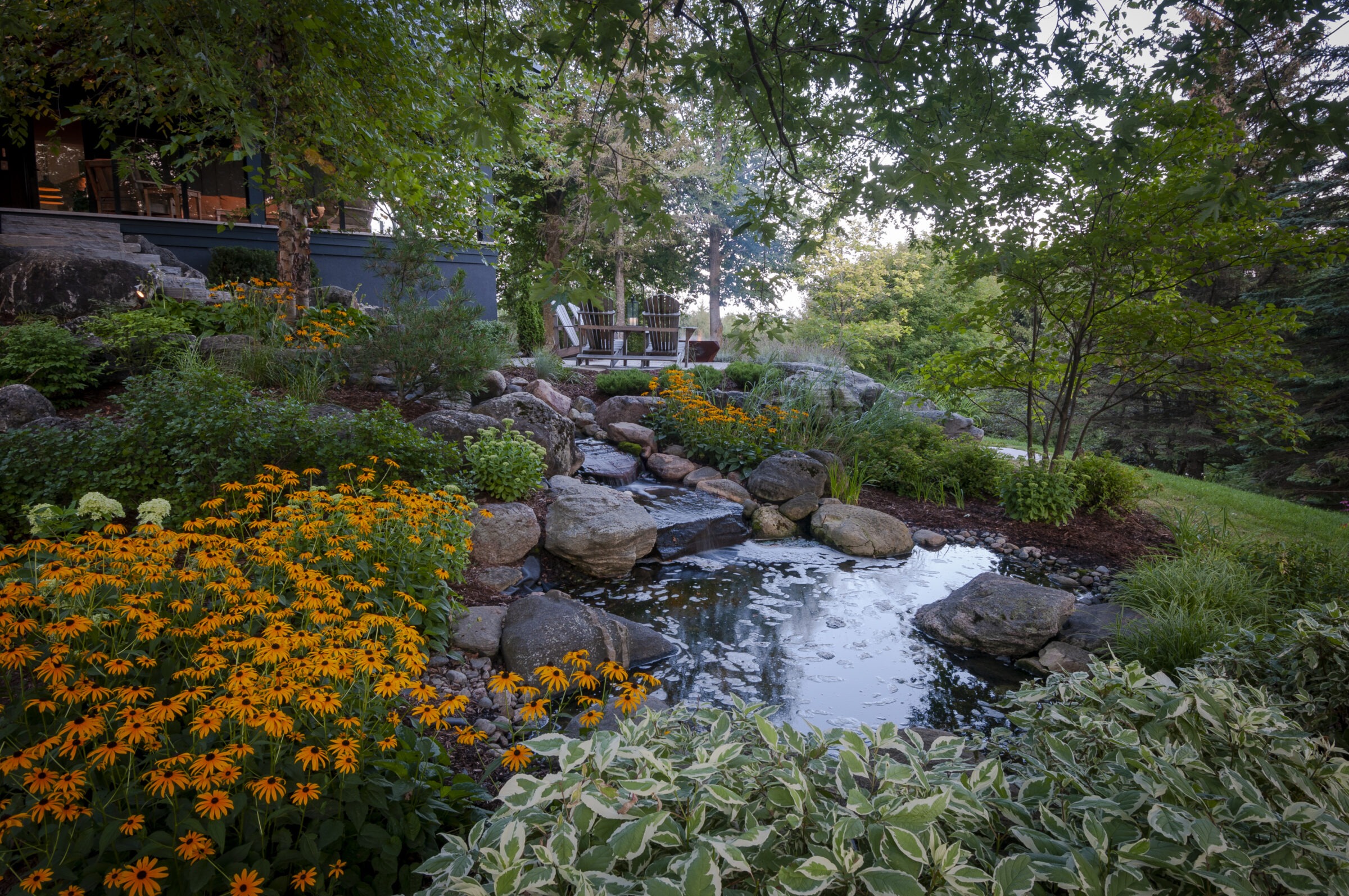 Lush garden scene with vibrant flowers, a small pond, and two chairs on a wooden deck, surrounded by trees under a clear sky.