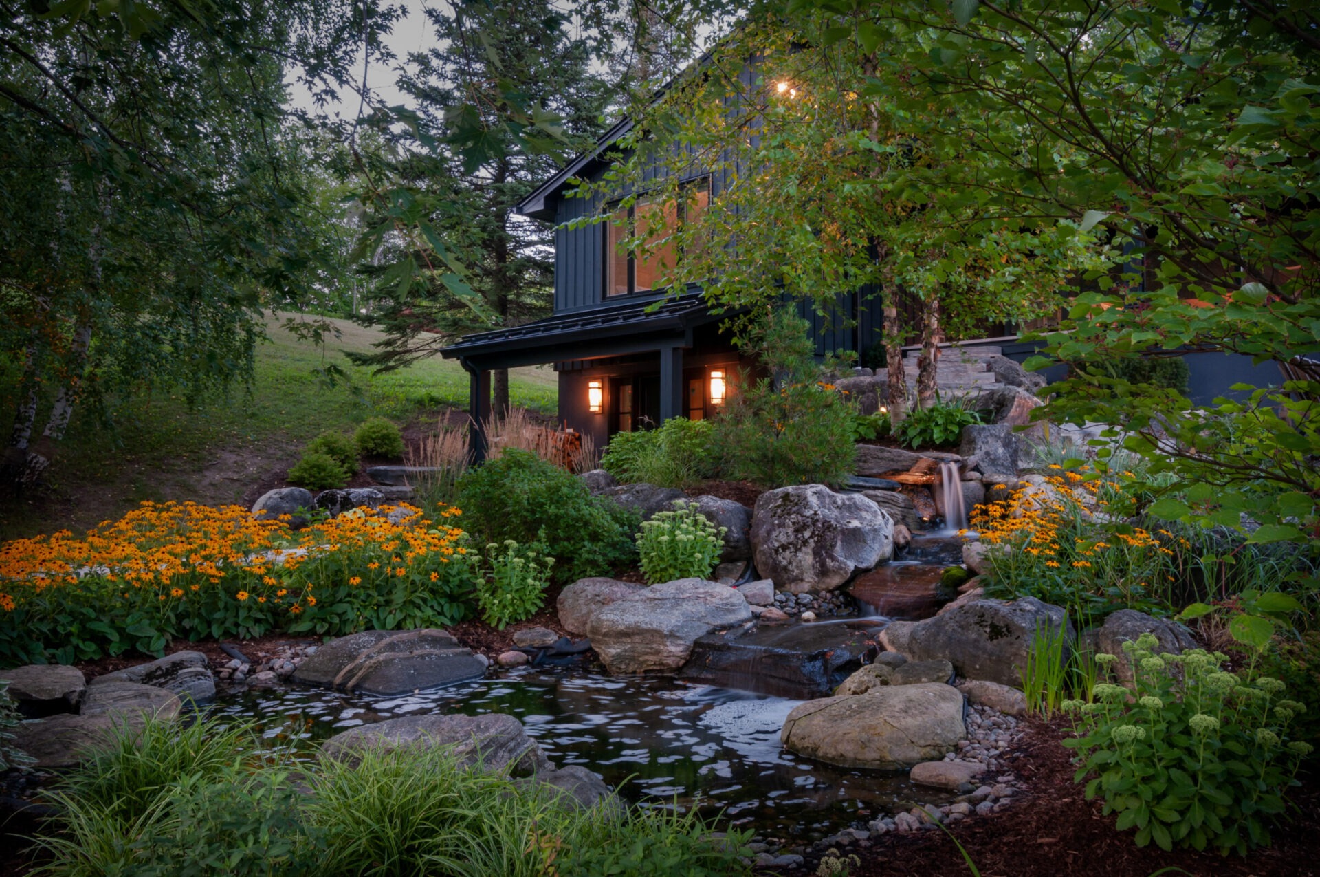 A cozy house is surrounded by lush greenery, vibrant flowers, rocks, and a serene pond with a small waterfall cascading gently.