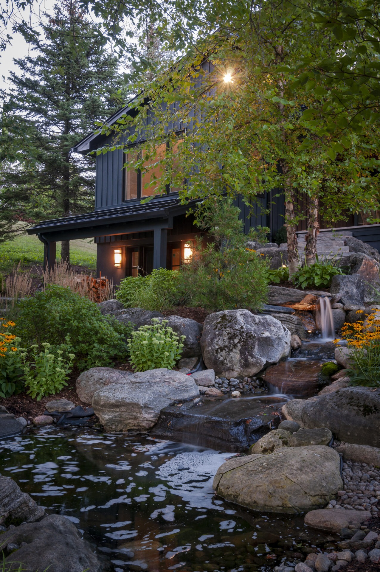Rustic house surrounded by lush greenery, rocks, and a small waterfall, creating a serene, natural setting with evening lights illuminating the cozy scene.