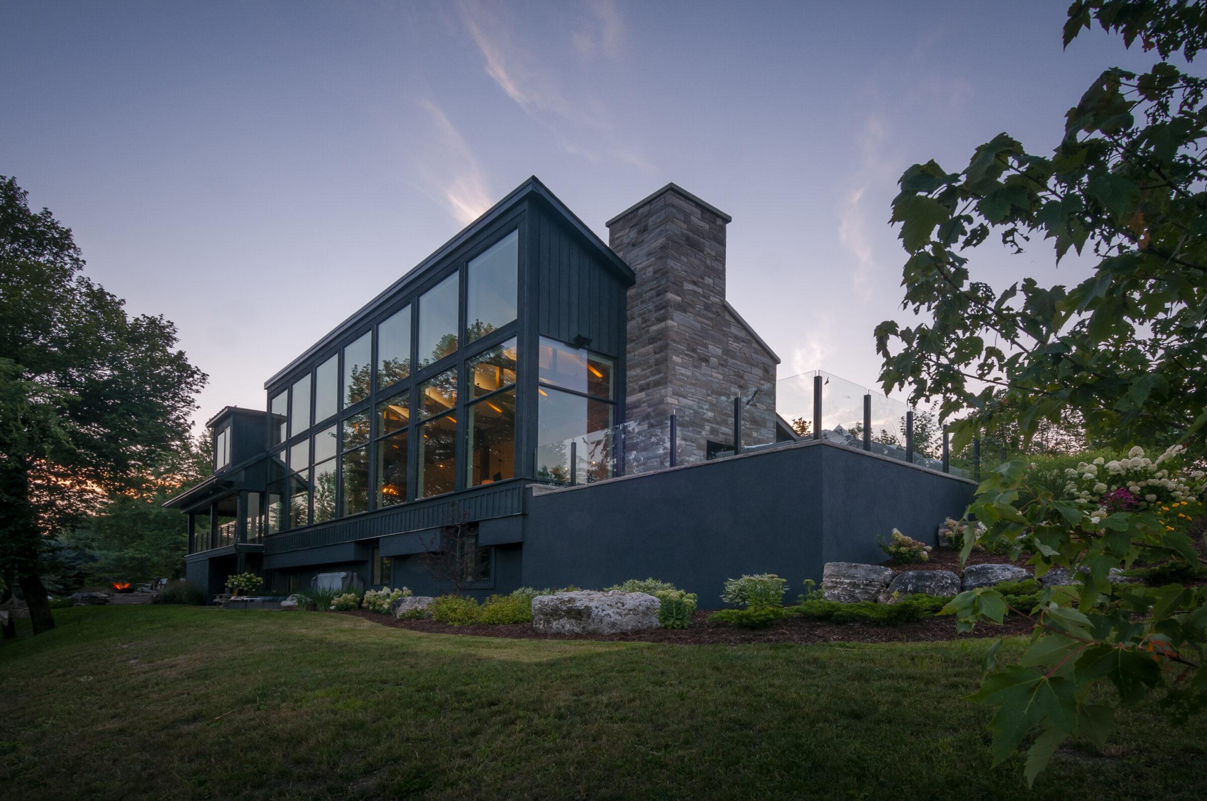 Modern house with large glass windows and stone chimney. Surrounded by trees and a well-maintained garden, under a twilight sky.