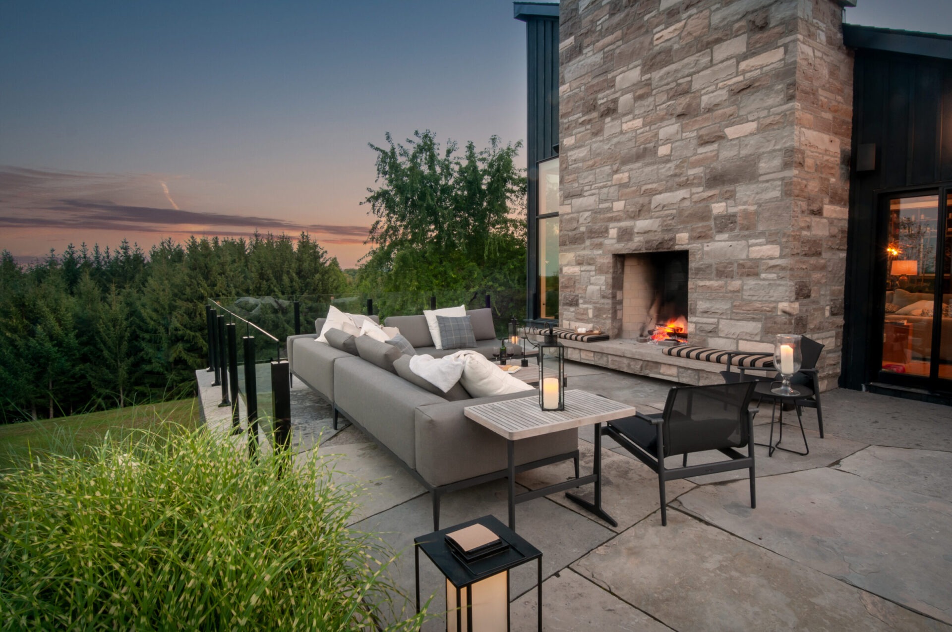Modern patio with stone fireplace and comfortable seating. Overlooks trees at sunset. Lanterns and cushions create a cozy atmosphere. No people present.