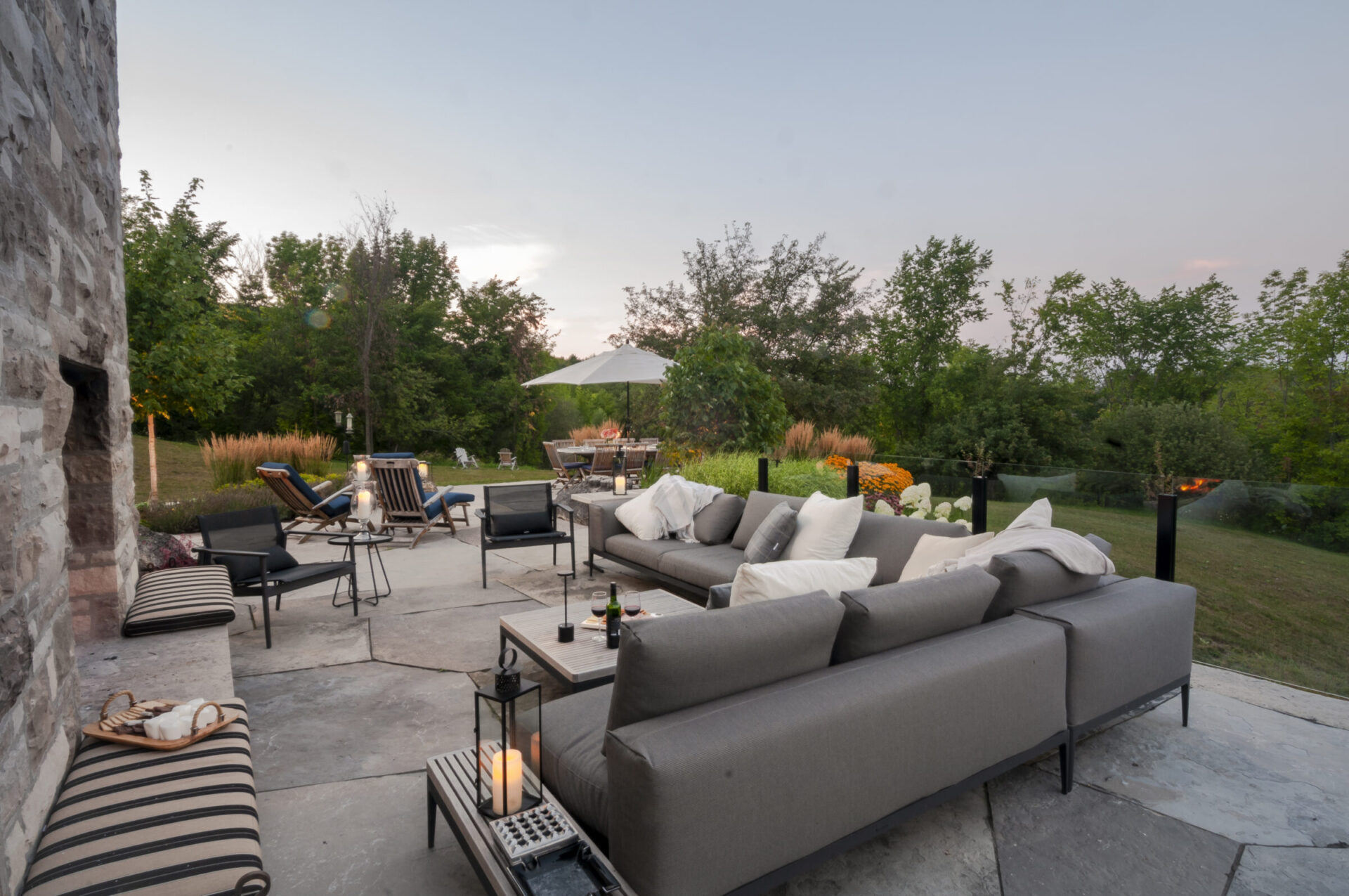 Outdoor patio with gray sofas, cushions, and lanterns. Surrounded by lush greenery and flowers; relaxing ambiance in natural setting at dusk.