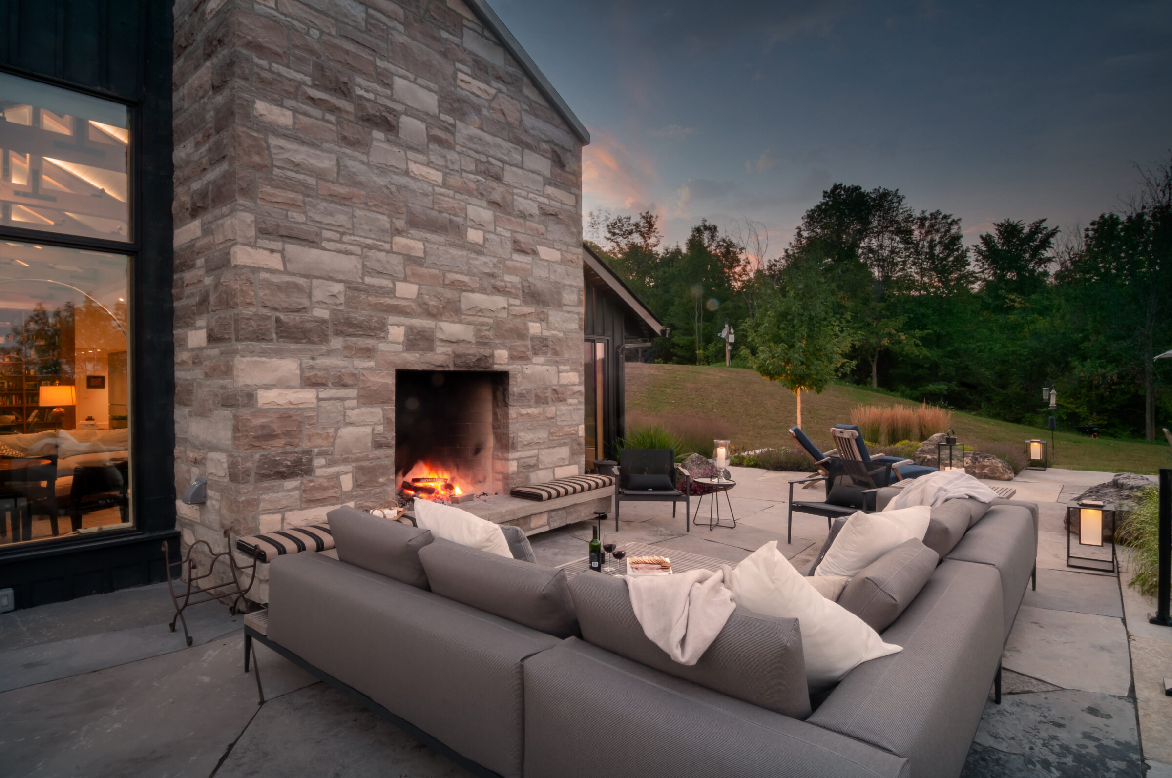 Cozy outdoor patio with a large stone fireplace, comfortable seating, and soft lighting, set against a twilight sky and lush greenery.