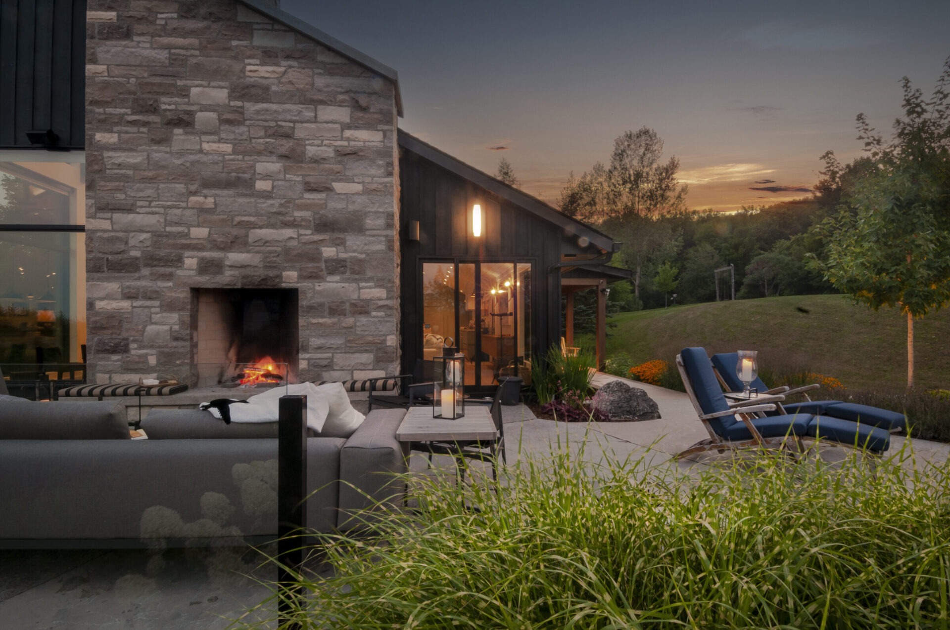 Modern house at sunset with stone fireplace, cozy patio furniture, and scenic greenery. Warm light enhances the serene outdoor atmosphere.