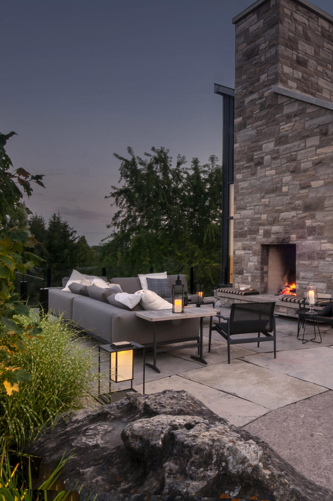 A cozy outdoor patio with a stone fireplace, cushioned seating, and lanterns set in a natural garden landscape under twilight skies.