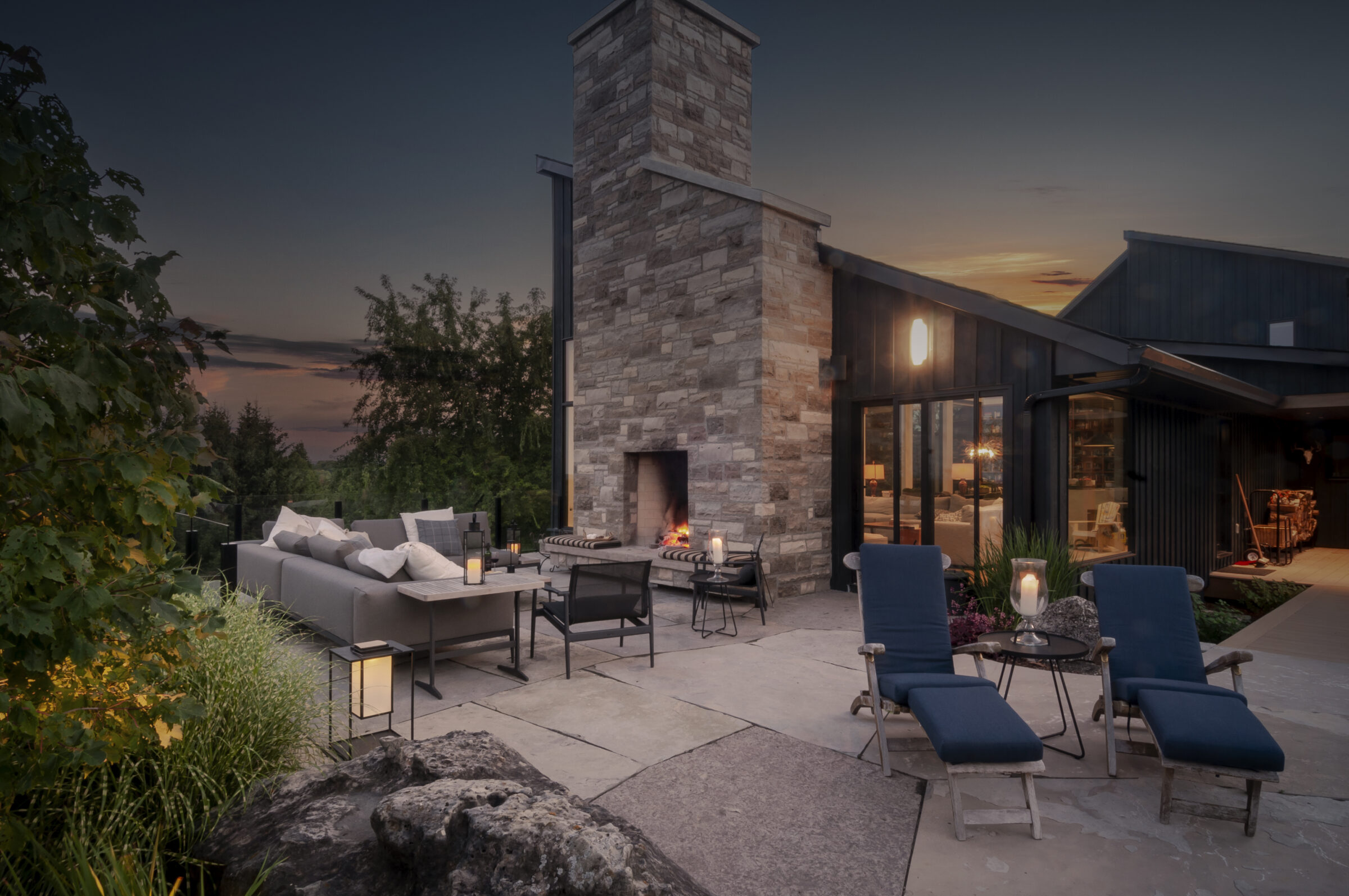 A stylish outdoor patio at sunset features a cozy seating area, stone fireplace, modern furniture, and ambient lighting surrounded by lush greenery.