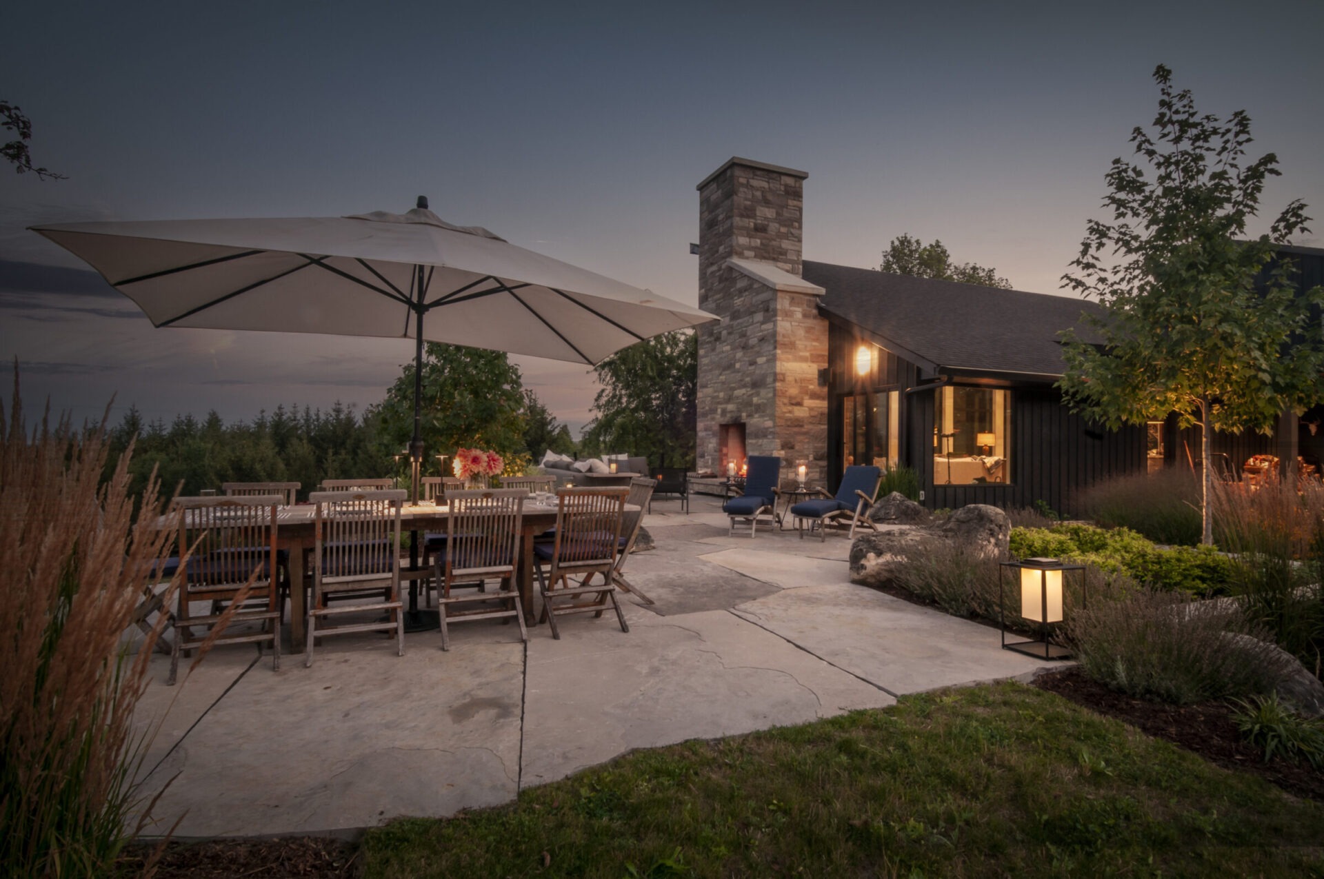 A serene patio scene at dusk features a large dining table under an umbrella, near a house with a stone chimney.