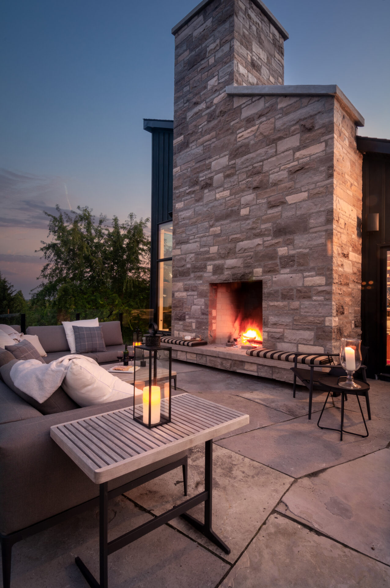 A cozy outdoor patio features a crackling fireplace, cushioned seating, and candlelit tables, nestled under a twilight sky near leafy trees.