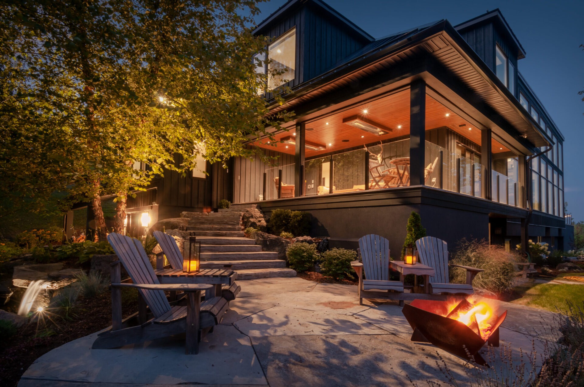 A modern house with an illuminated patio, Adirondack chairs, fire pit, and scenic landscaping under glowing evening lights and lush trees.