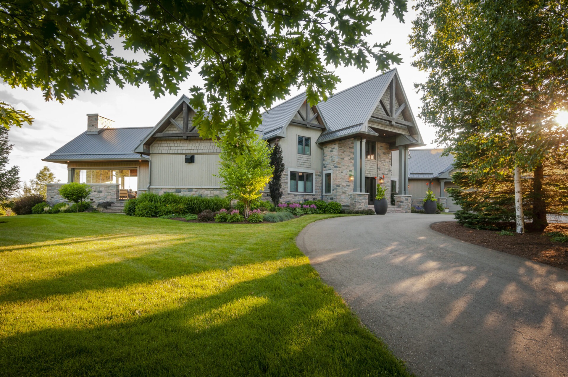 A modern stone house with a metal roof is surrounded by a well-manicured lawn, trees, and a curving driveway under a clear sky.