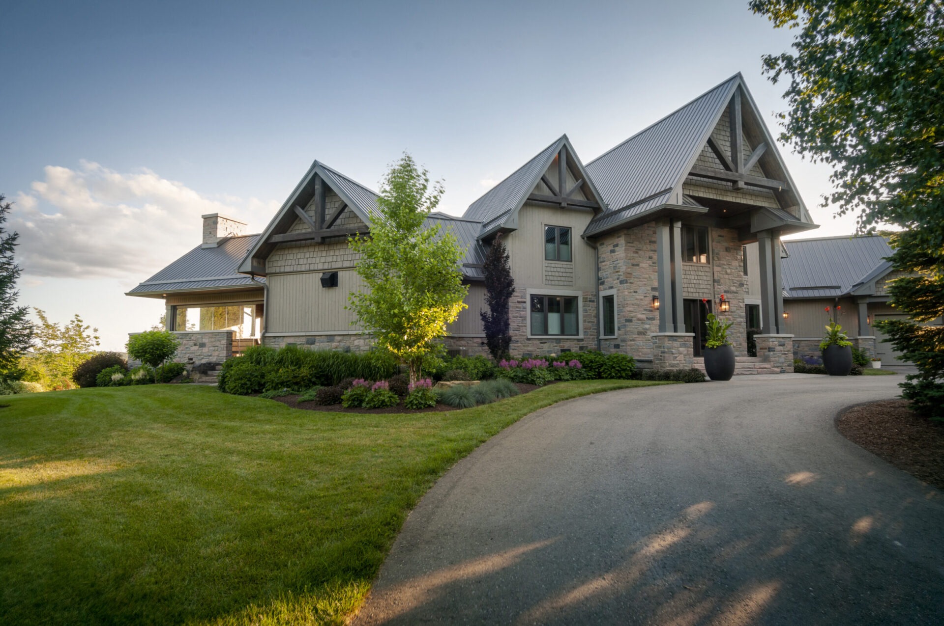 Large modern house with stone and wood accents, surrounded by a well-maintained garden and trees, under a clear blue sky.