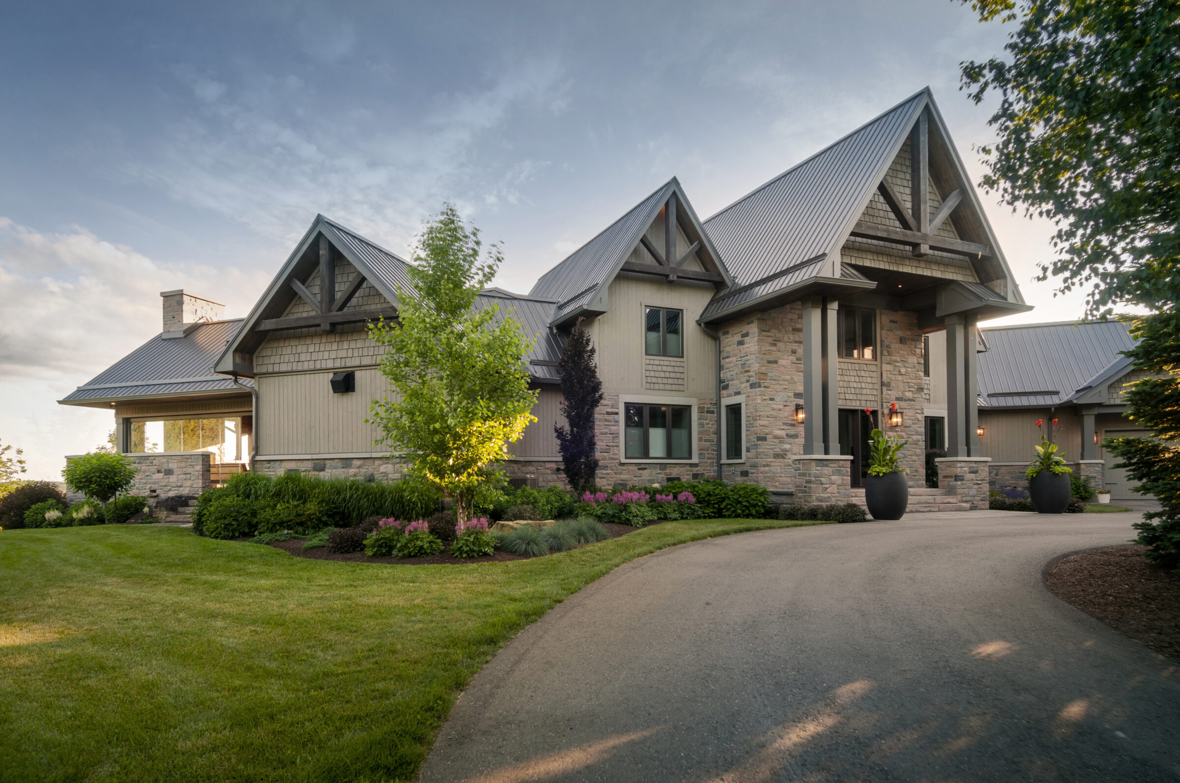 A modern house with stone and wood elements, surrounded by a lush garden and curved driveway under a clear sky.