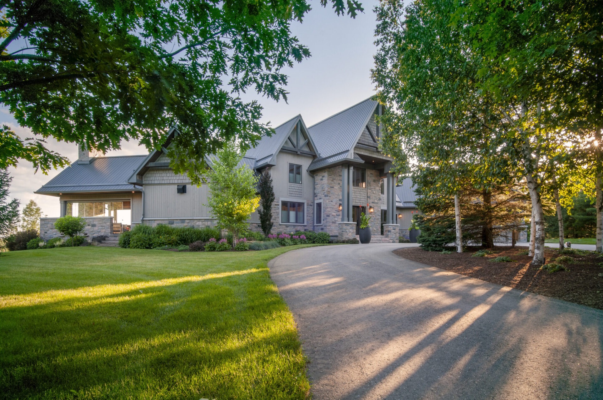A modern stone and wood house with a sloped metal roof, surrounded by lush greenery and a winding driveway in a serene setting.