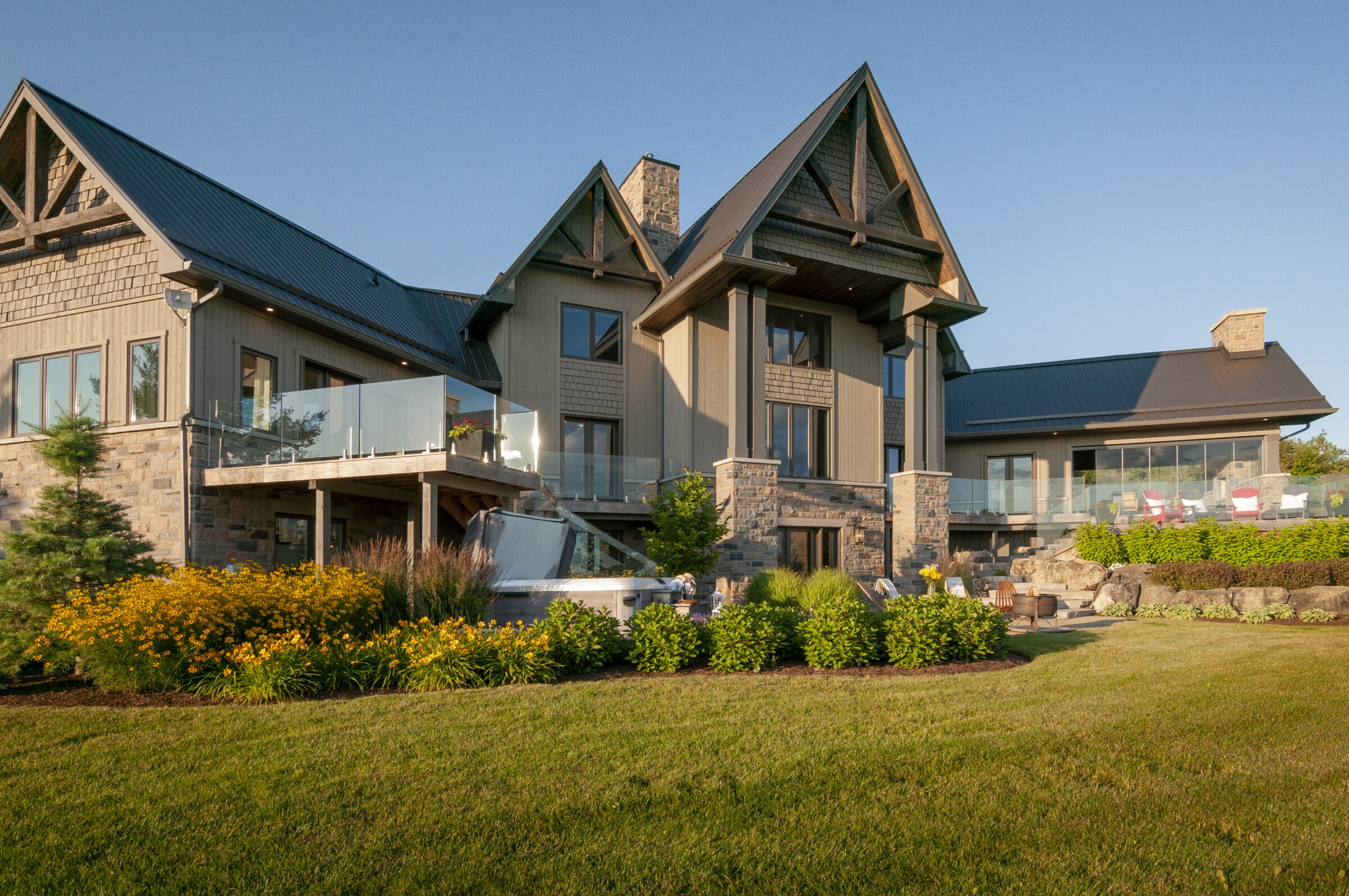 A large modern house with wooden and stone features, spacious glass balconies, surrounded by manicured lawns and vibrant flower gardens.