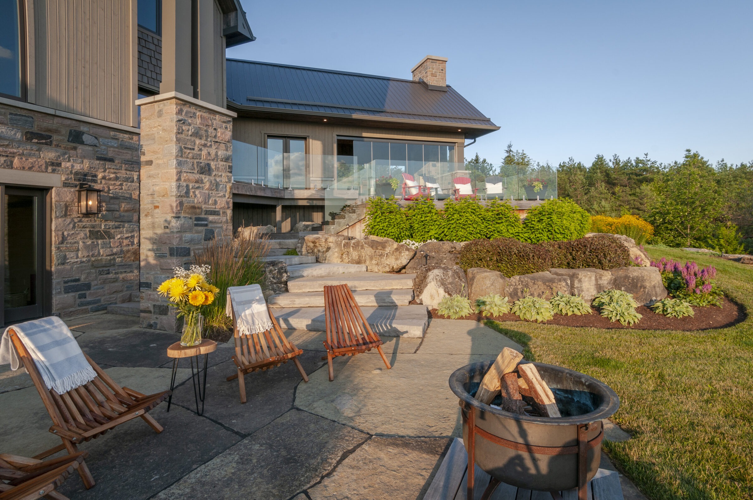 Contemporary stone house with patio, wooden chairs, and fire pit. Lush landscaping and glass railing enhance the scenic, tranquil outdoor space.