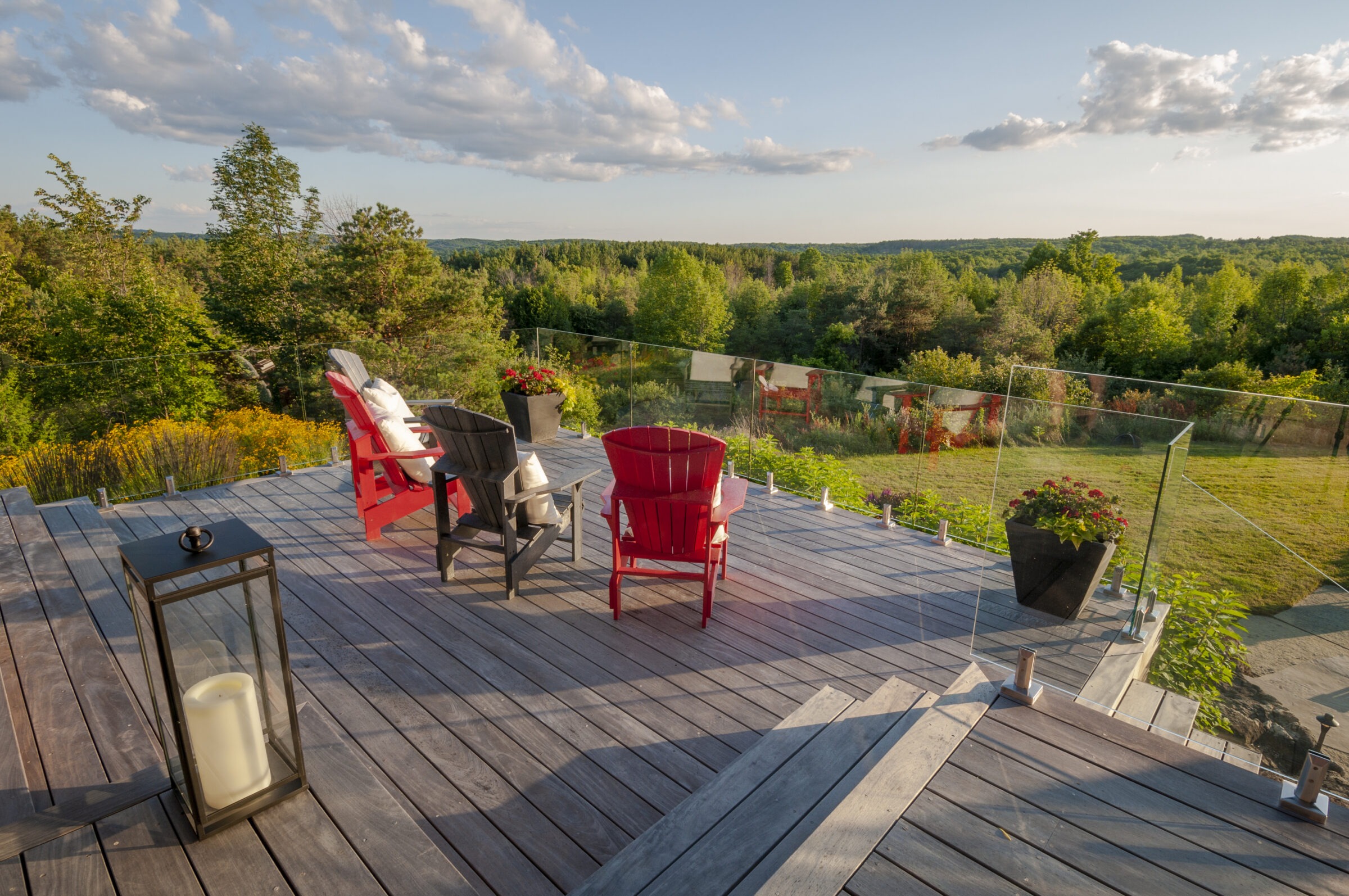 Wooden deck overlooking lush green landscape, featuring colorful chairs and potted plants, framed by glass railing under a blue sky. Peaceful outdoor setting.