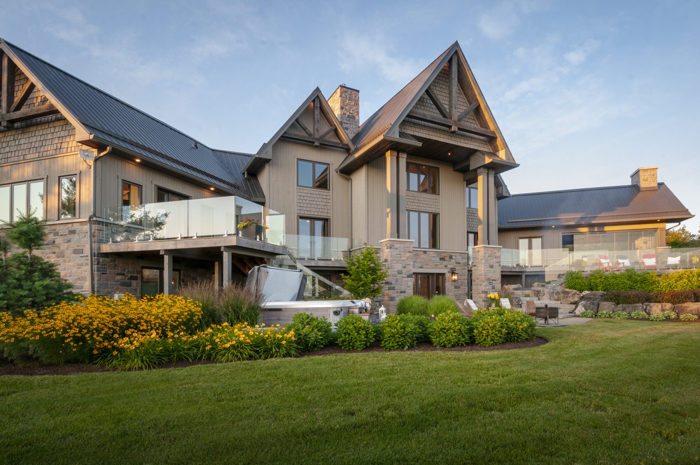 A modern house with stone and wood accents features a glass balcony, surrounded by lush landscaping and a spacious green lawn.