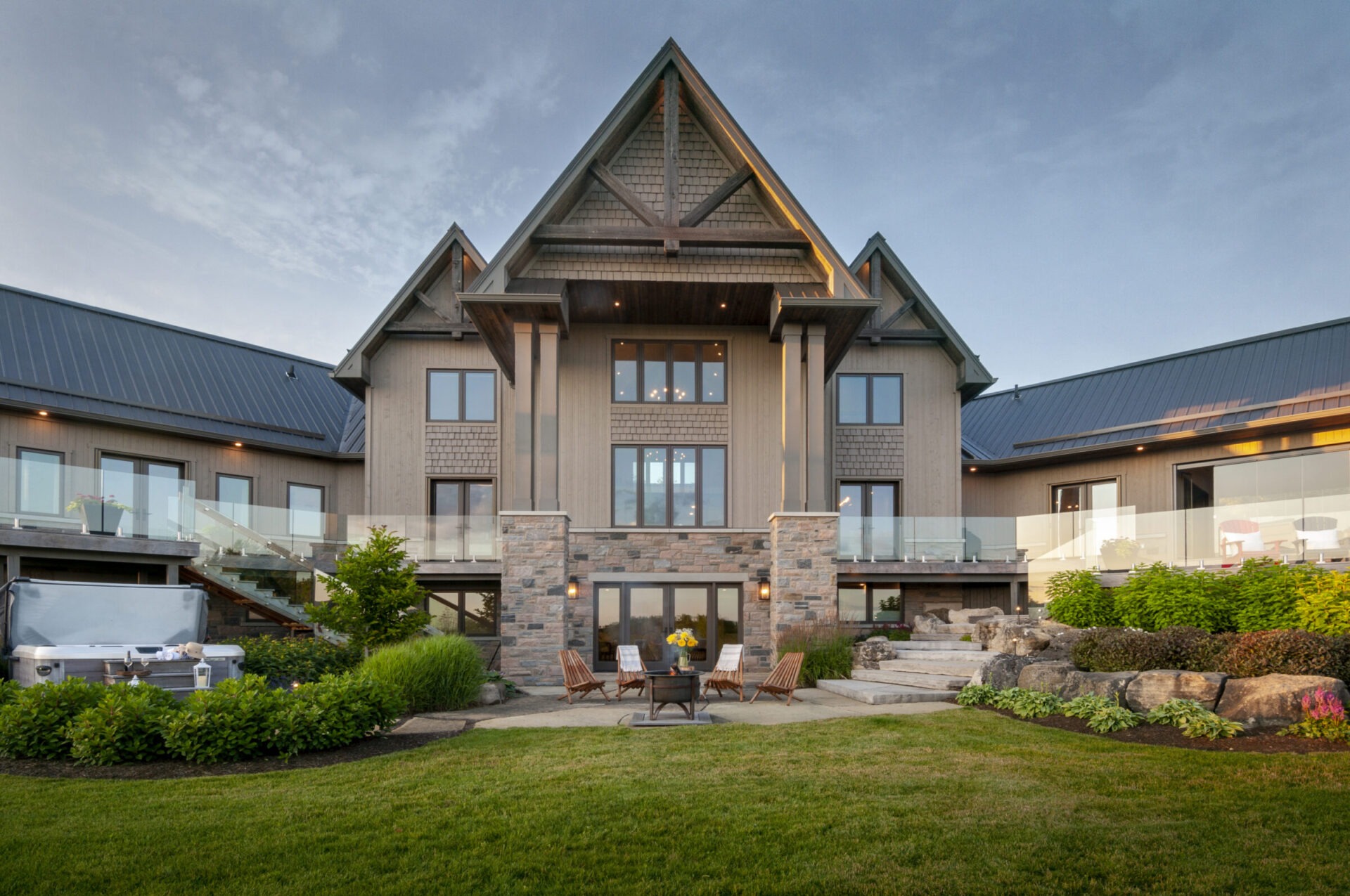 A modern, large house with wooden and stone accents, surrounded by greenery and a patio area with chairs and a hot tub.