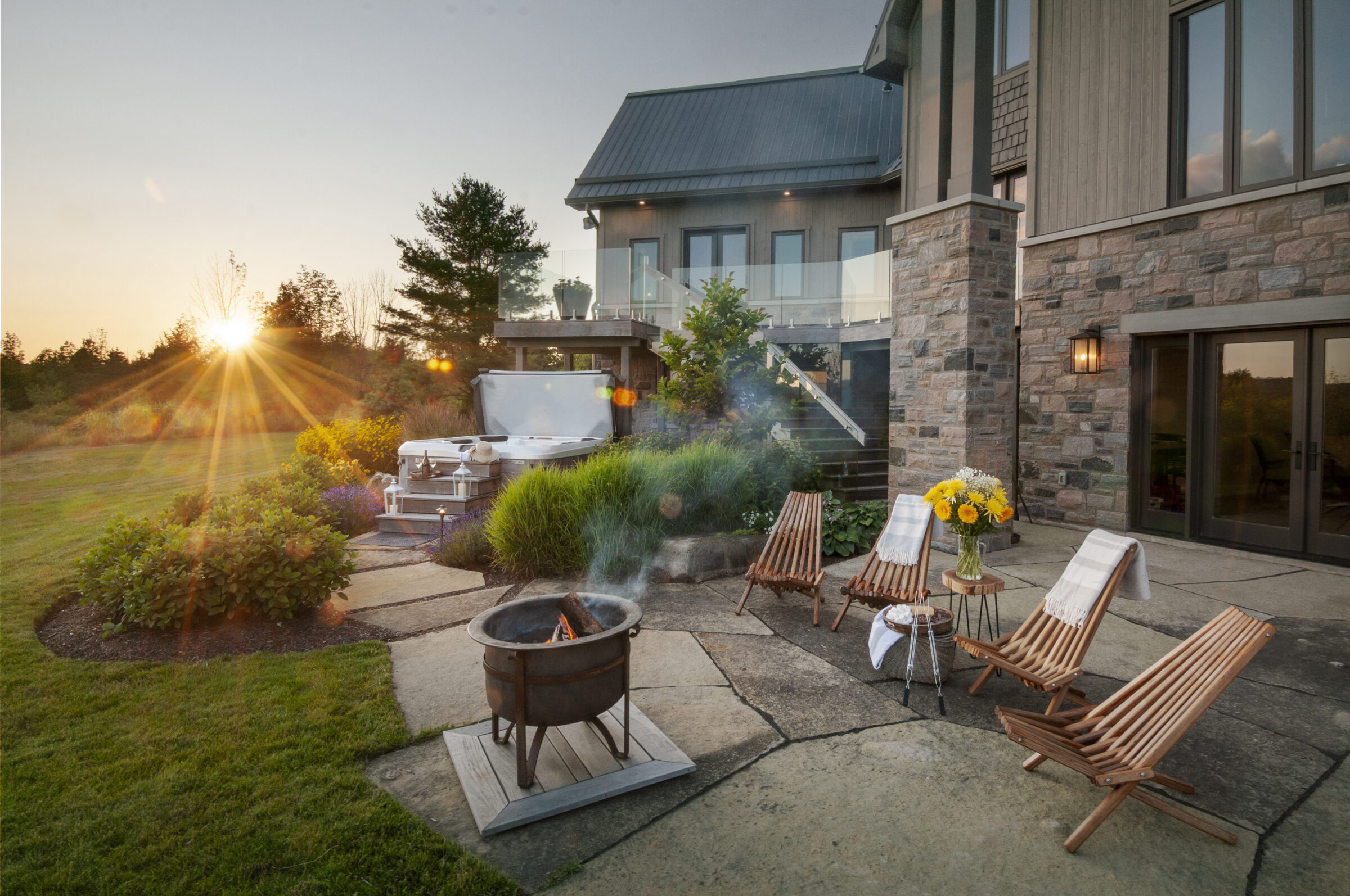 A serene backyard scene with a patio, fire pit, chairs, flowers, and sunset over a lush garden beside a modern house.
