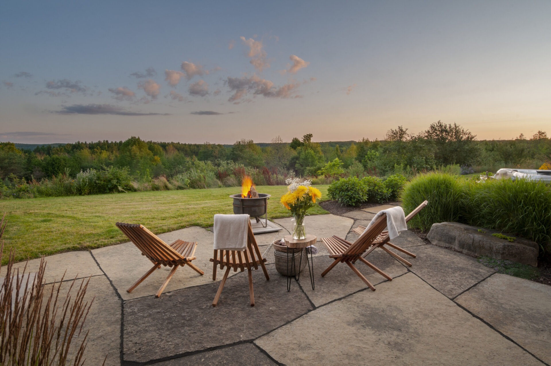 Four wooden chairs surround a fire pit on a patio, overlooking a lush green landscape and colorful sky during sunset.