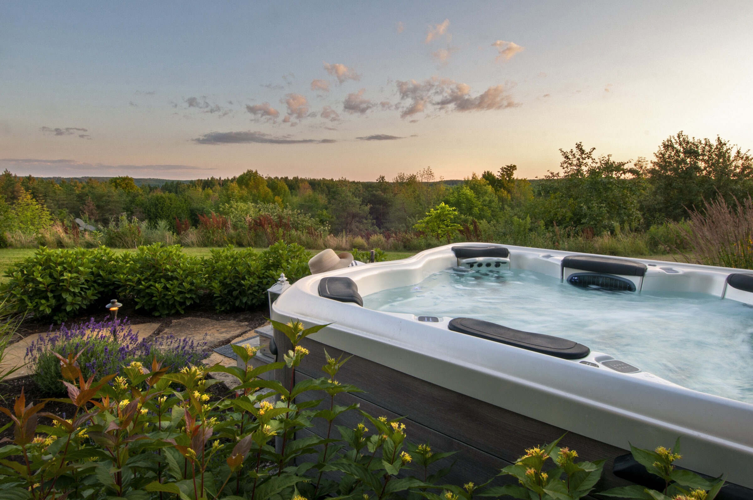 A hot tub with a hat rests in a lush garden at sunset, surrounded by vibrant greenery and flowers.