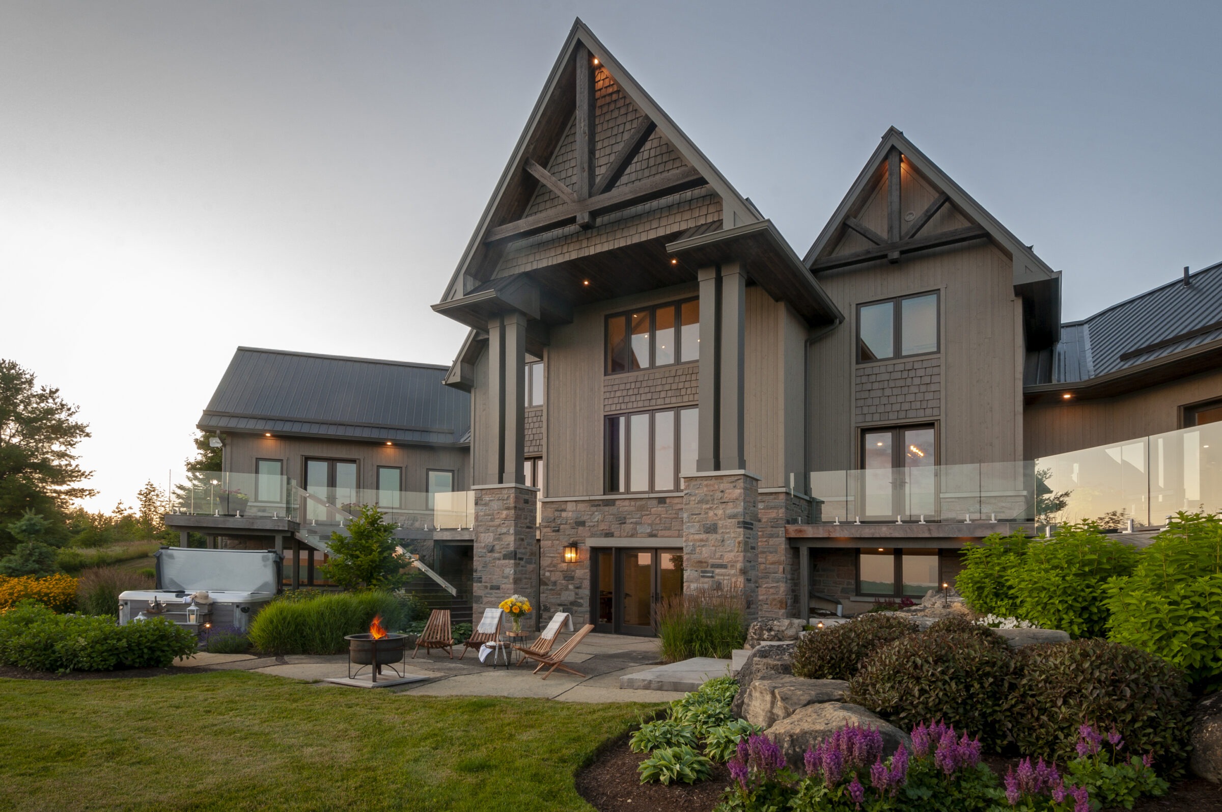 A modern, rustic house with large windows, stone accents, and manicured garden chairs around a fire pit at sunset.