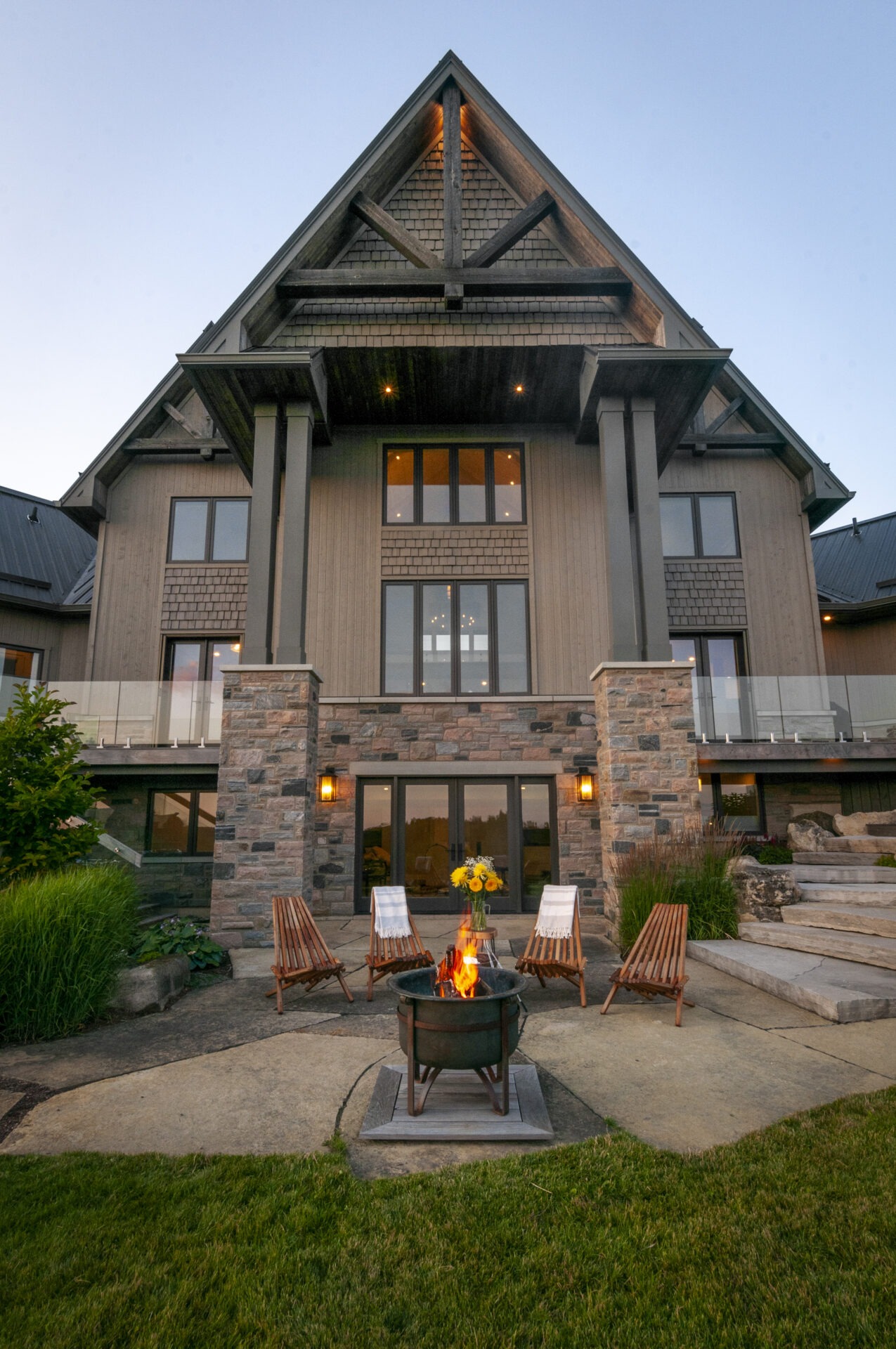 Large modern house with stone and wood exterior, surrounded by lawn and patio. Chairs circle a fire pit, adding warmth and ambiance.