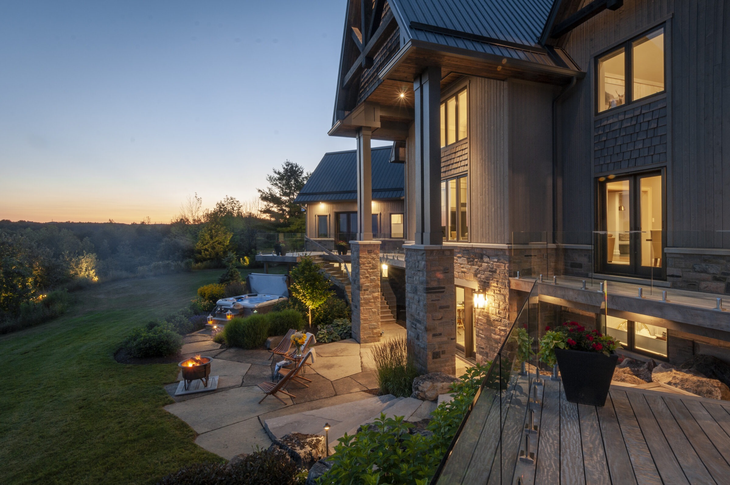 Modern house at dusk with large windows, wooden deck, patio, hot tub, and fire pit, surrounded by trees and a manicured lawn.