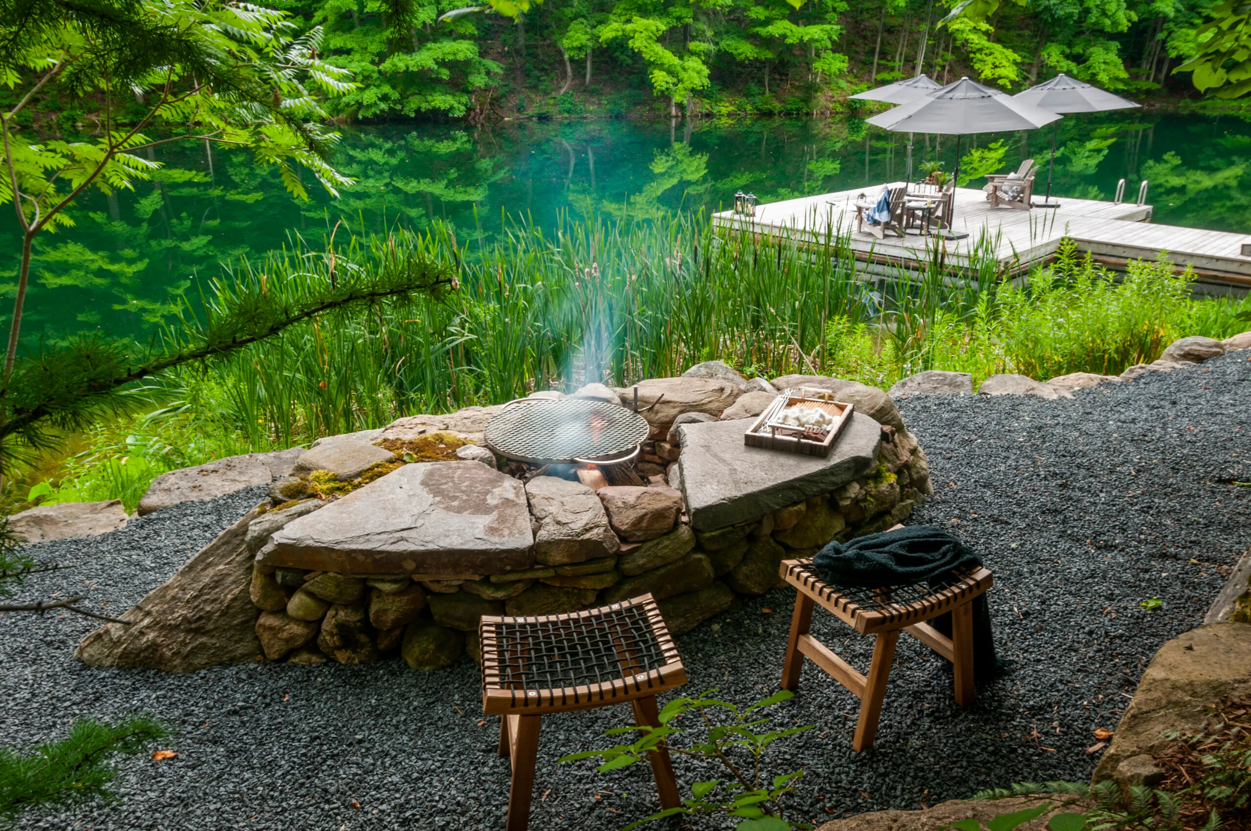 A serene lakeside scene with a stone fire pit, chairs, dock, and lush greenery reflecting in calm water under umbrellas.