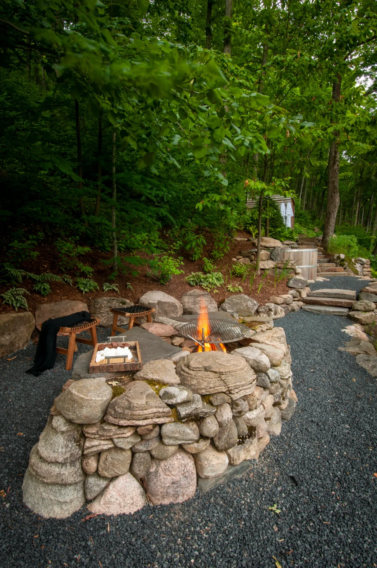 A natural stone fire pit with burning logs, surrounded by chairs, marshmallows, and lush greenery, creates a serene outdoor retreat.