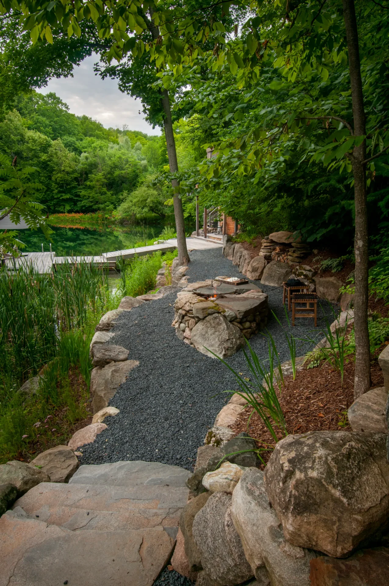 A serene garden pathway with stone steps and lush greenery leads to a small pond. Dense trees surround the tranquil setting.