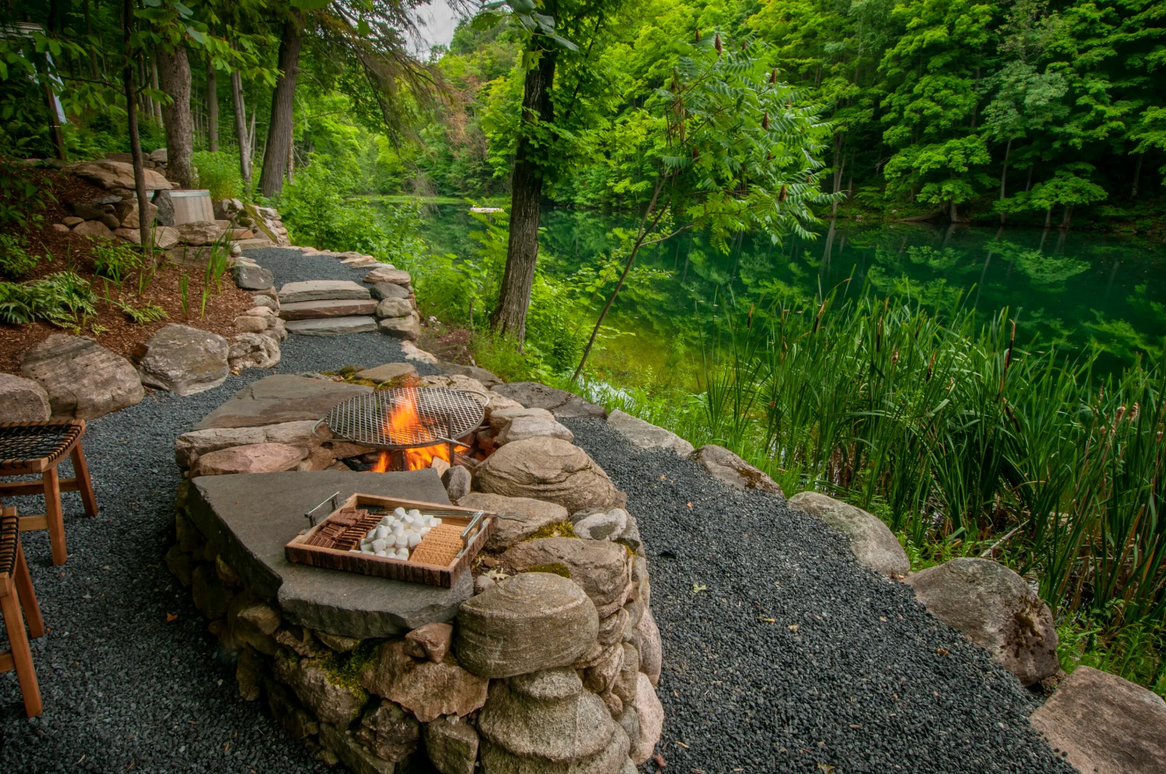 A serene forest setting by a lake, with a stone fire pit, grill, and s'mores ingredients on a stone slab.