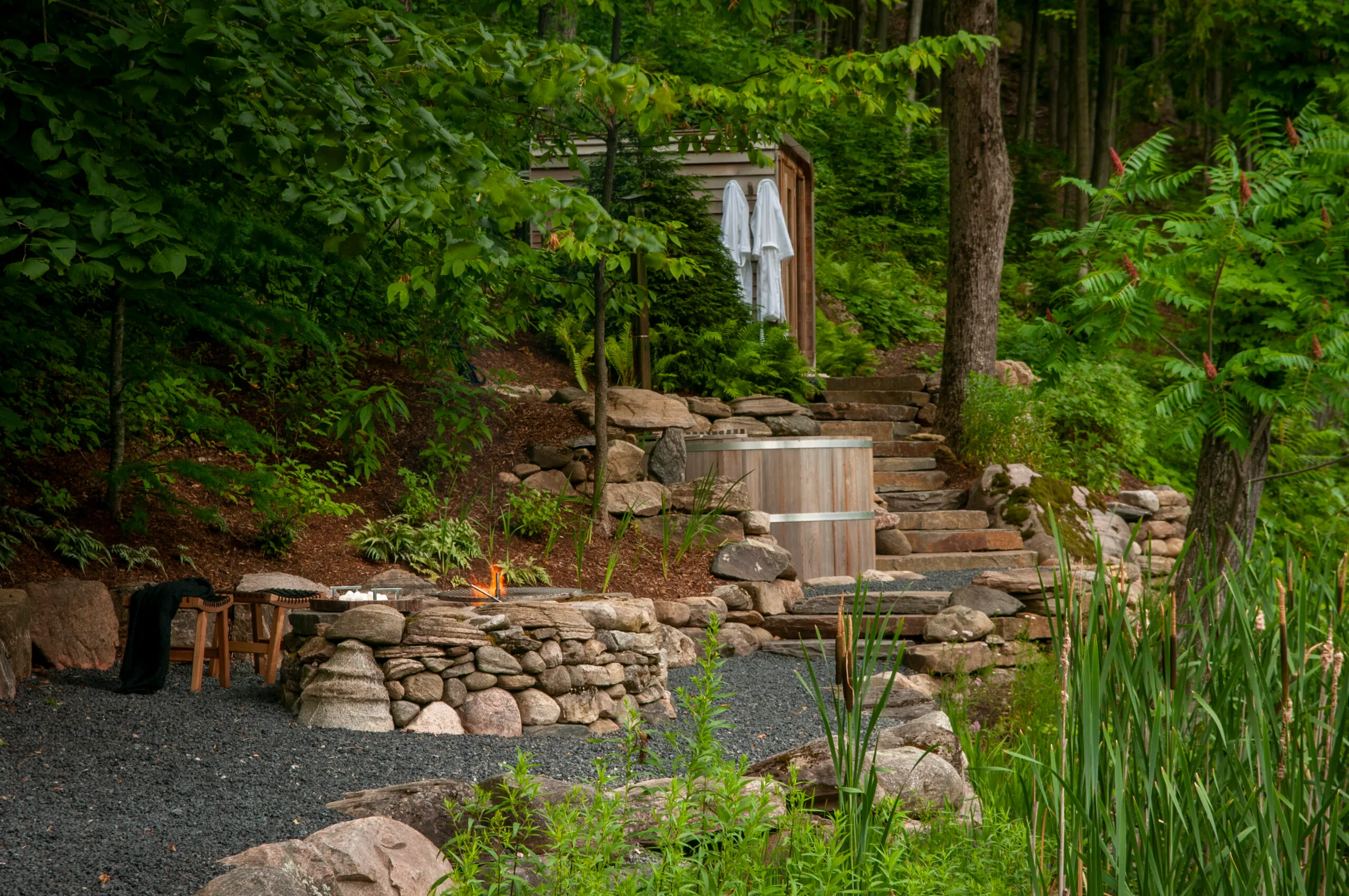 A serene forest setting features a stone fire pit, rustic seating, wooden hot tub, and stairs leading to a quaint cabin retreat.
