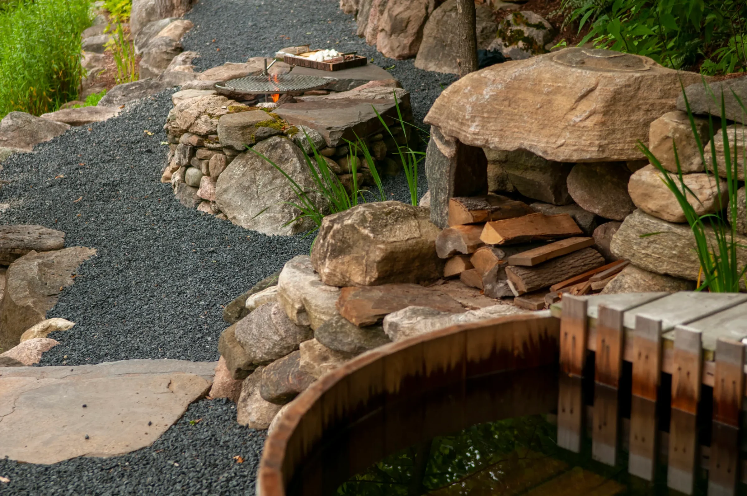 Stone pathway with a rustic fire pit, stacked wood, and a wooden-topped hot tub, surrounded by greenery and natural rocks.