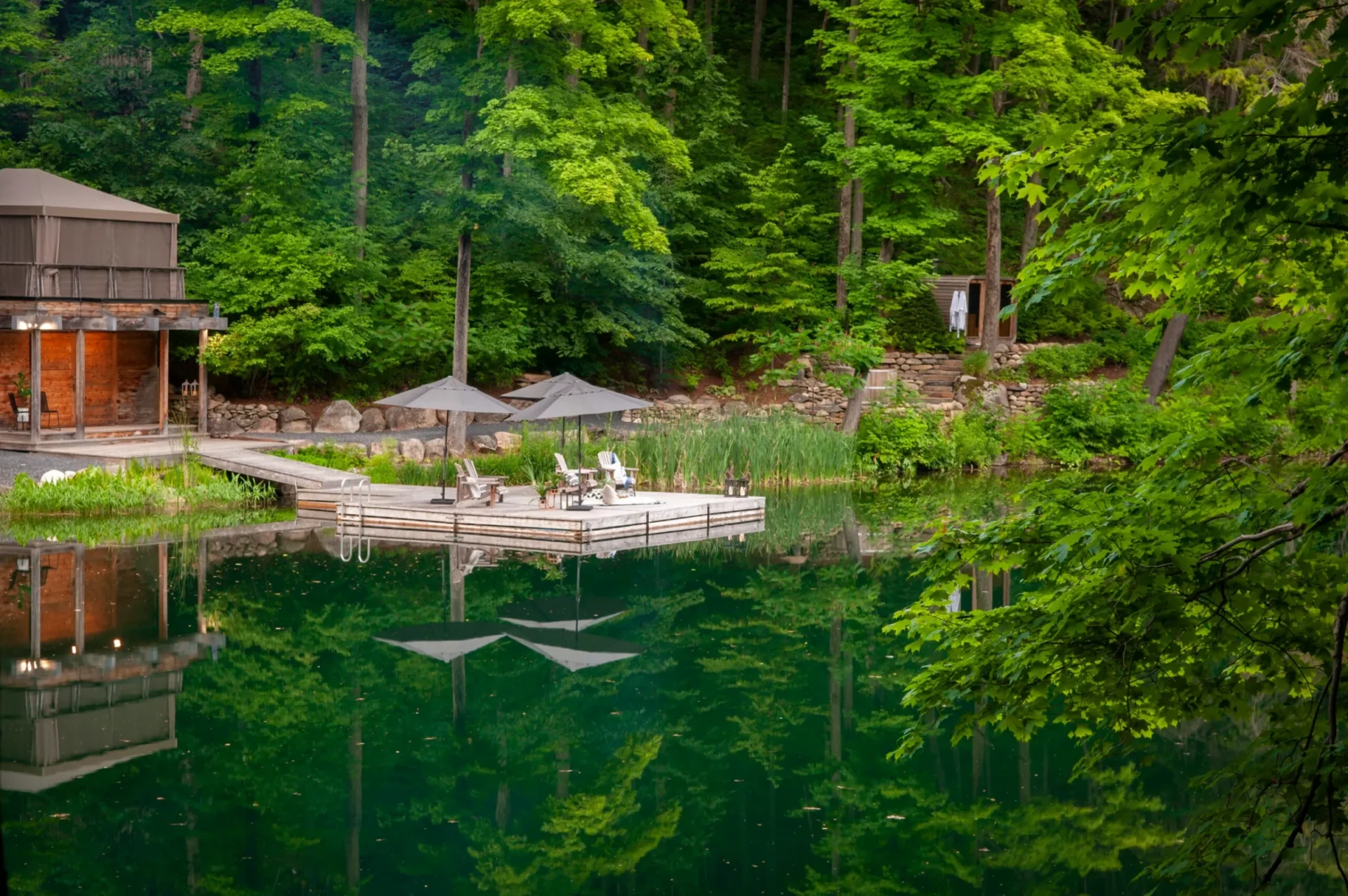 A serene forest setting with a wooden deck by a reflective lake, featuring chairs, umbrellas, and lush greenery surrounding a wooden building.